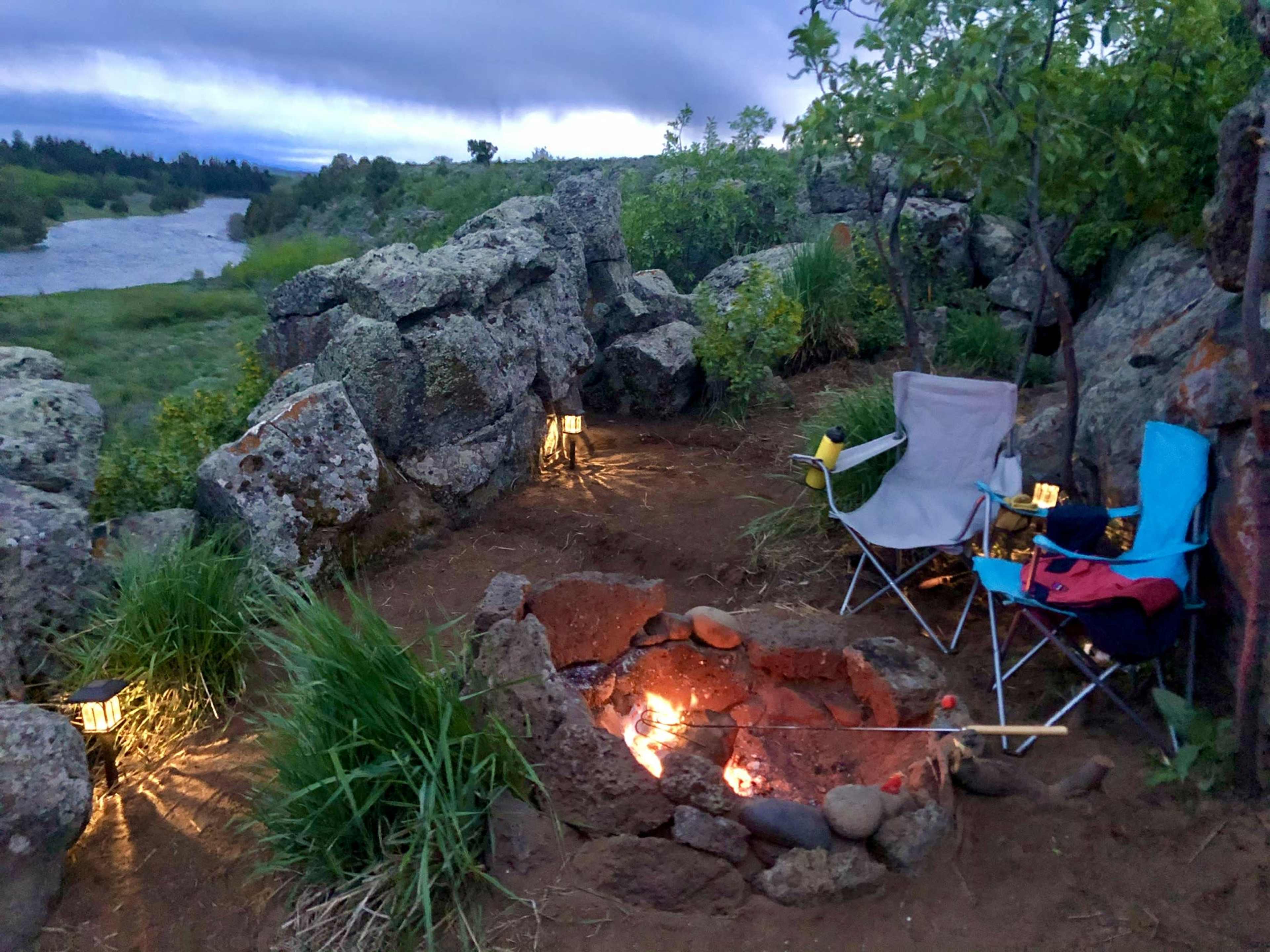 A small, rocky campsite features a fire pit surrounded by two chairs and lanterns, with a river and greenery in the background.