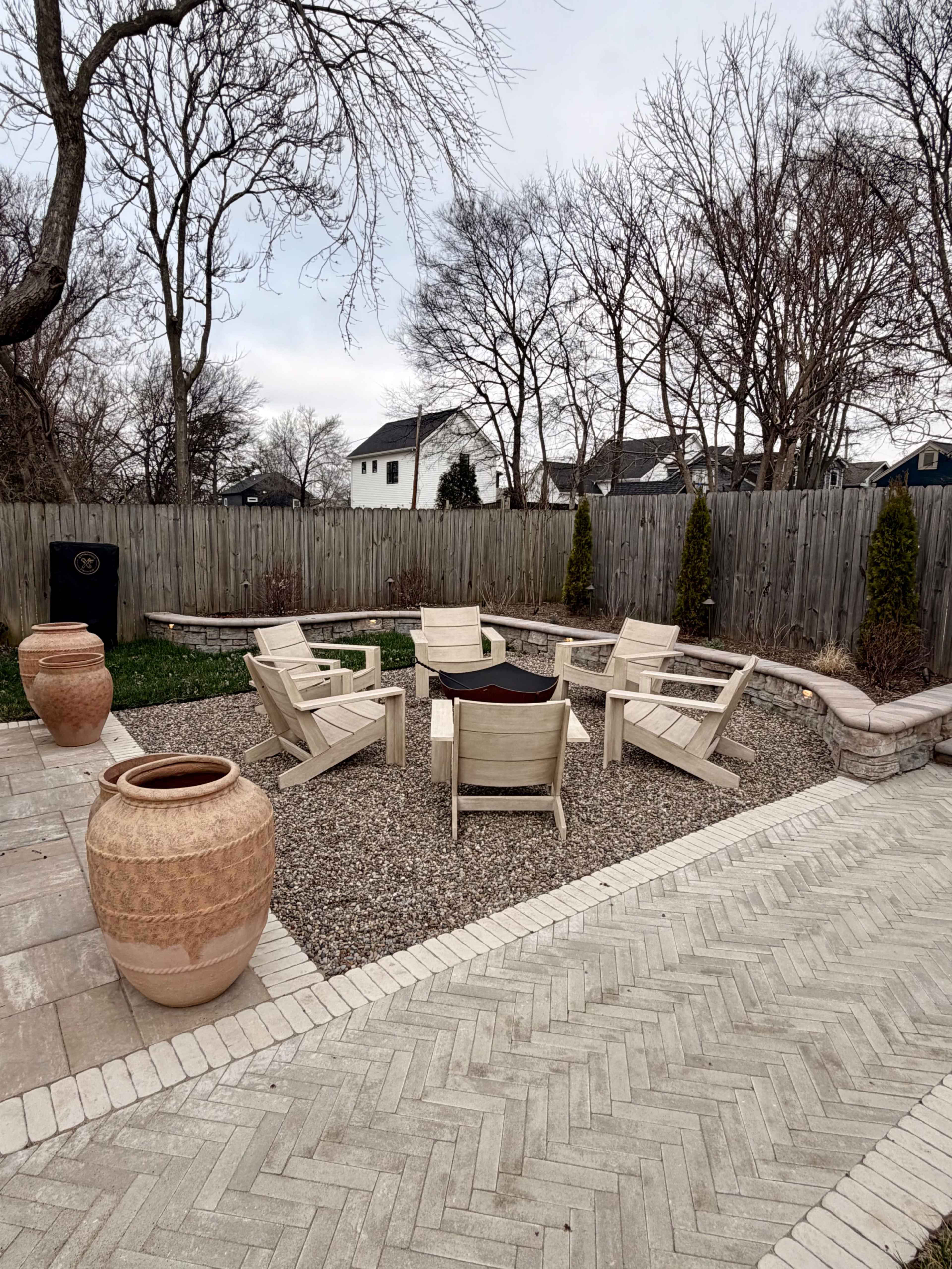 The image shows a circular seating area surrounded by gravel, featuring wooden chairs arranged around a central table, with large clay pots positioned nearby in a residential backyard.