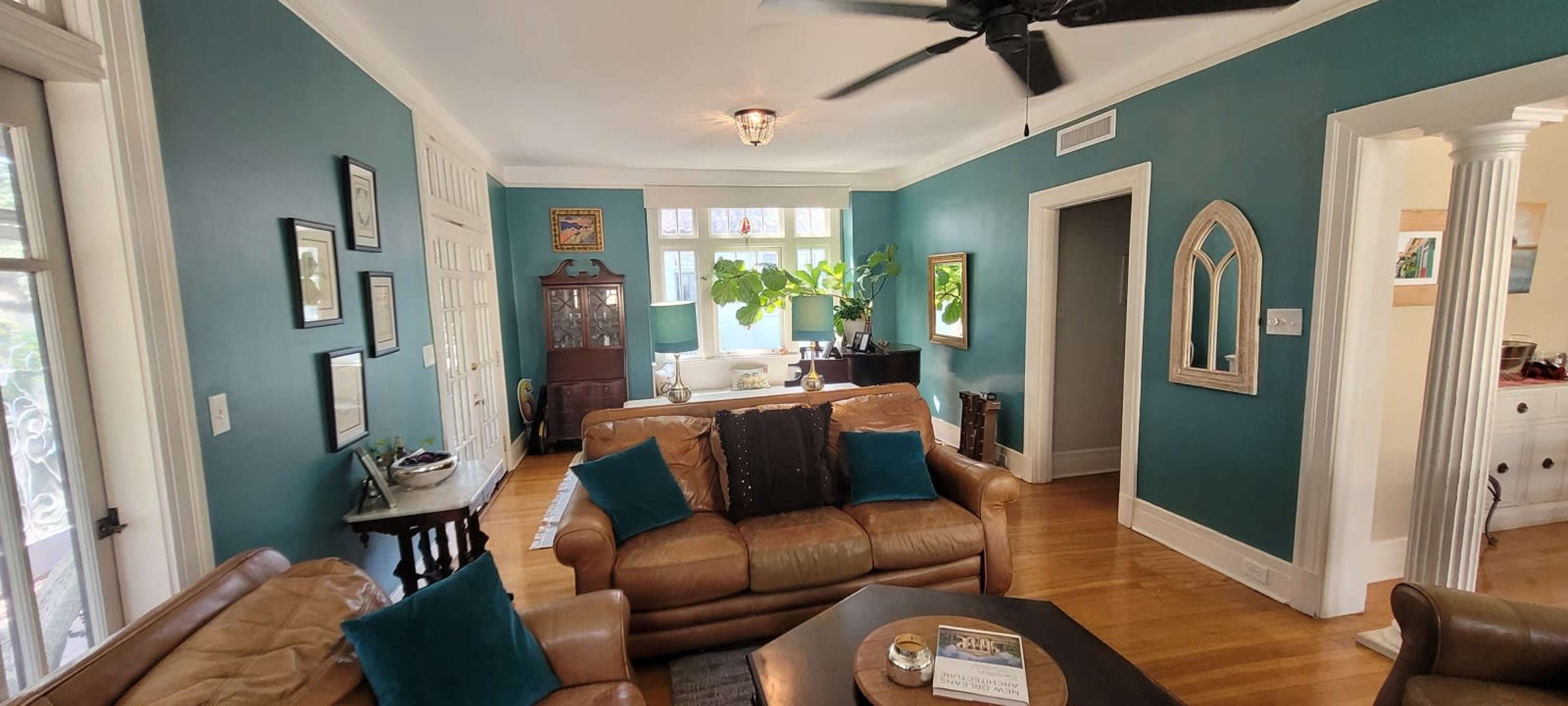 The image shows a living room with teal walls, featuring a mix of leather and fabric seating, a coffee table, and a large window allowing natural light.