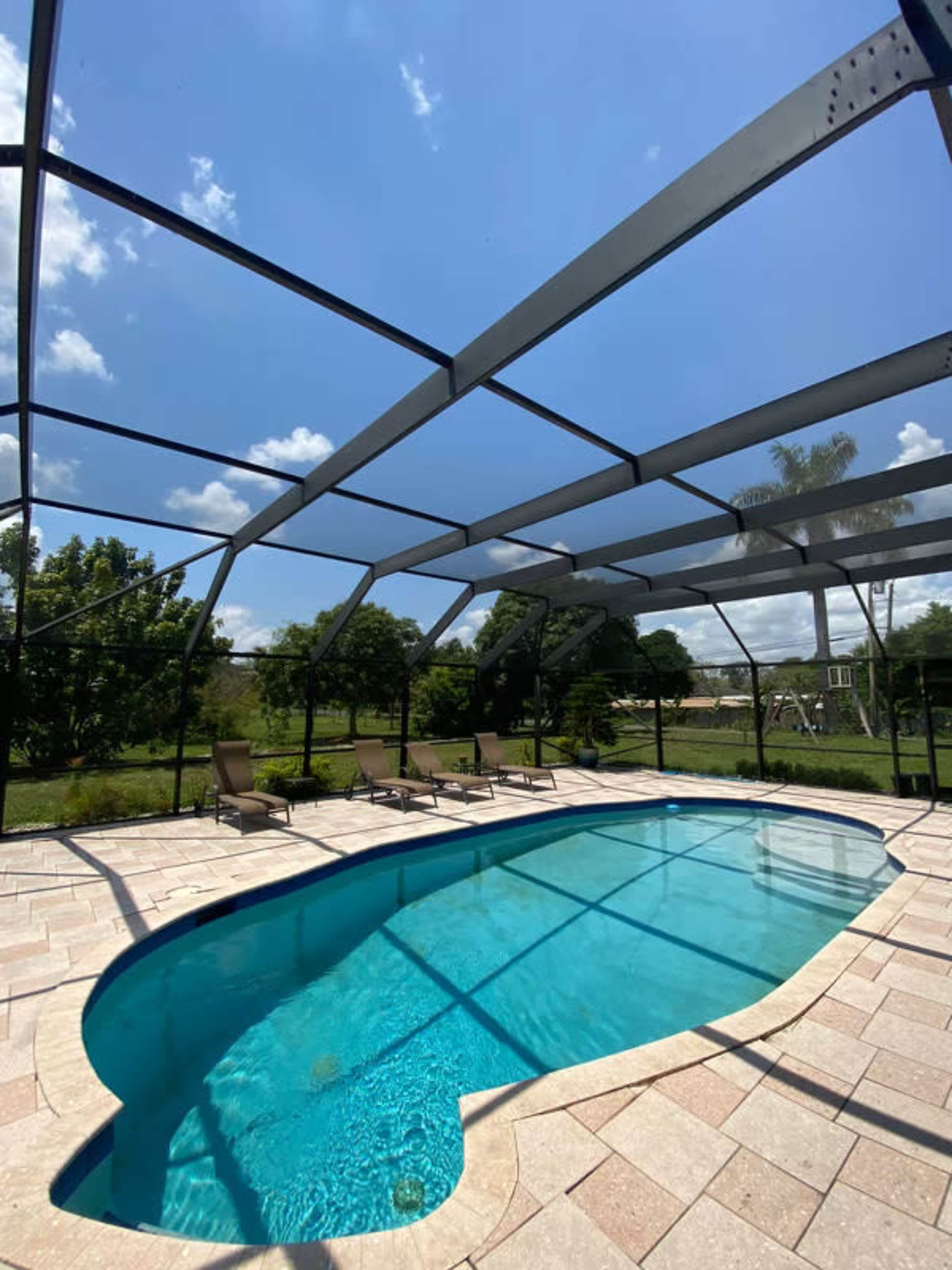 A rectangular pool surrounded by a patio with lounge chairs, enclosed by a screen, and set against a backdrop of greenery and blue sky.