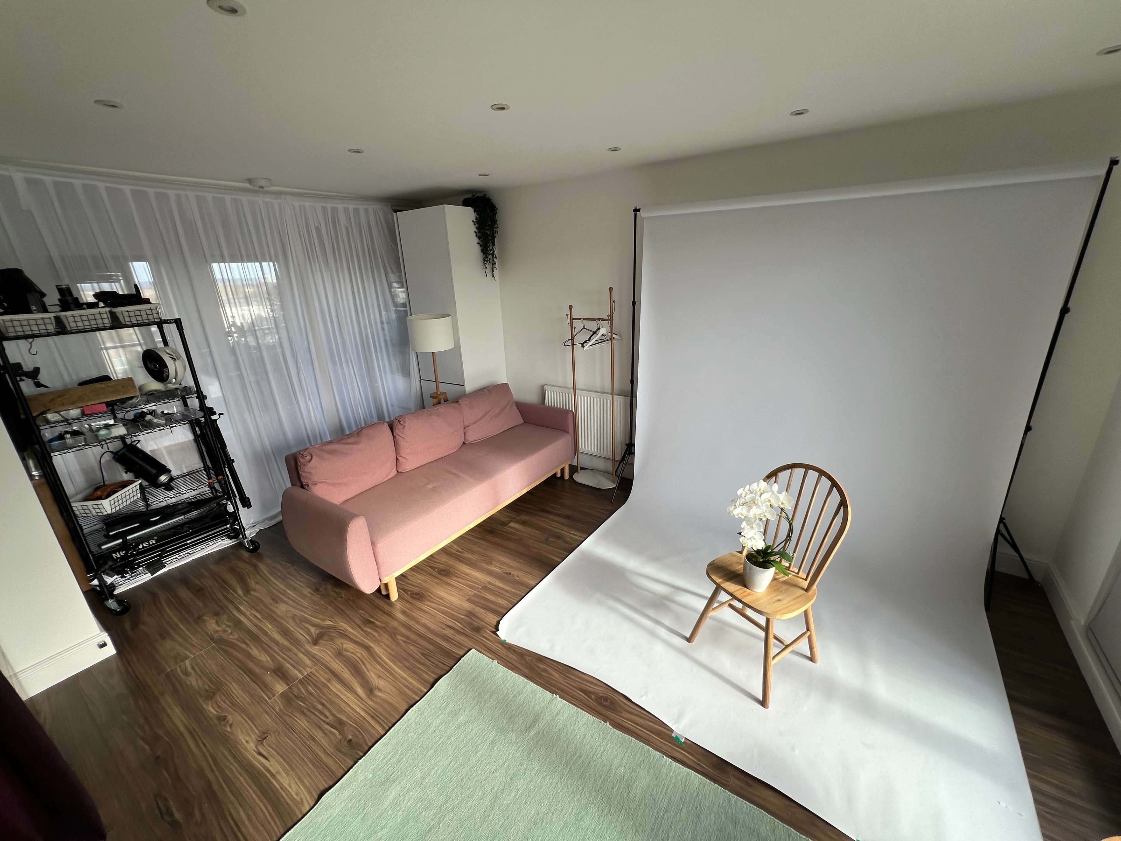 The image shows a bright room with a pink sofa, a wooden chair on a white backdrop, and a small table with flowers.