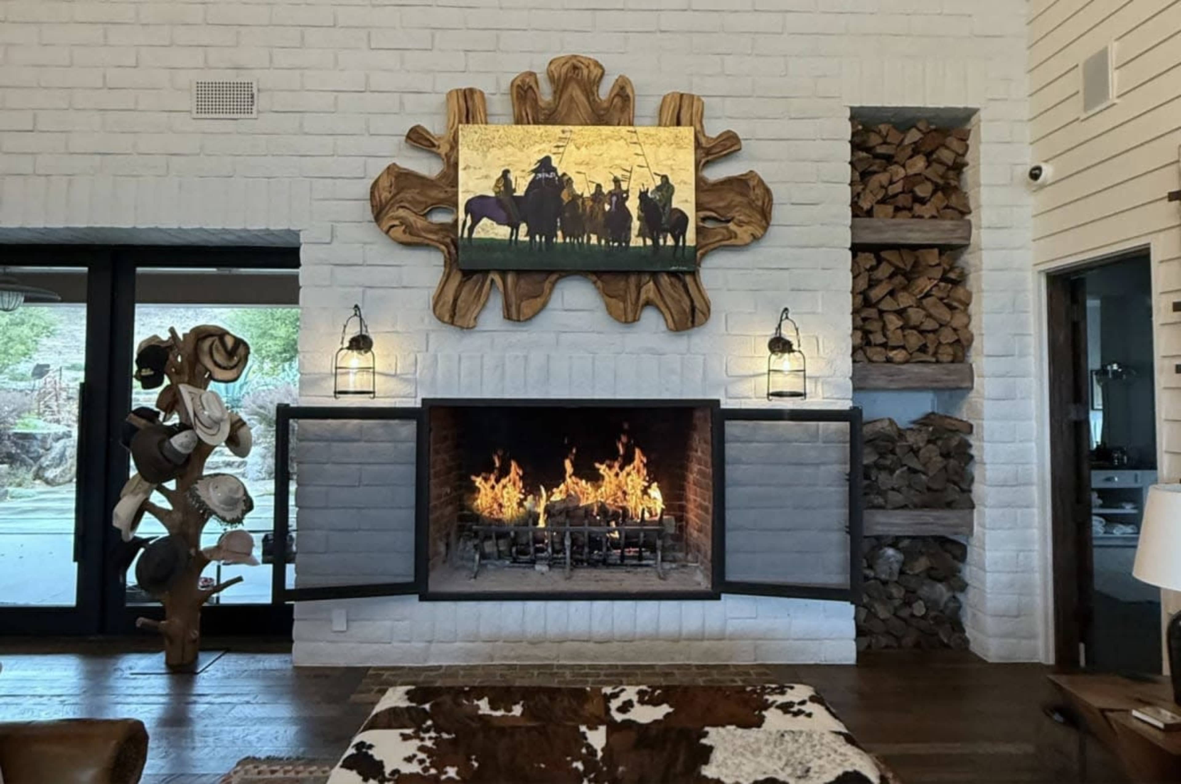 The image shows a modern living room with a central fireplace, a large piece of artwork above it, and neatly stacked firewood on the right side.