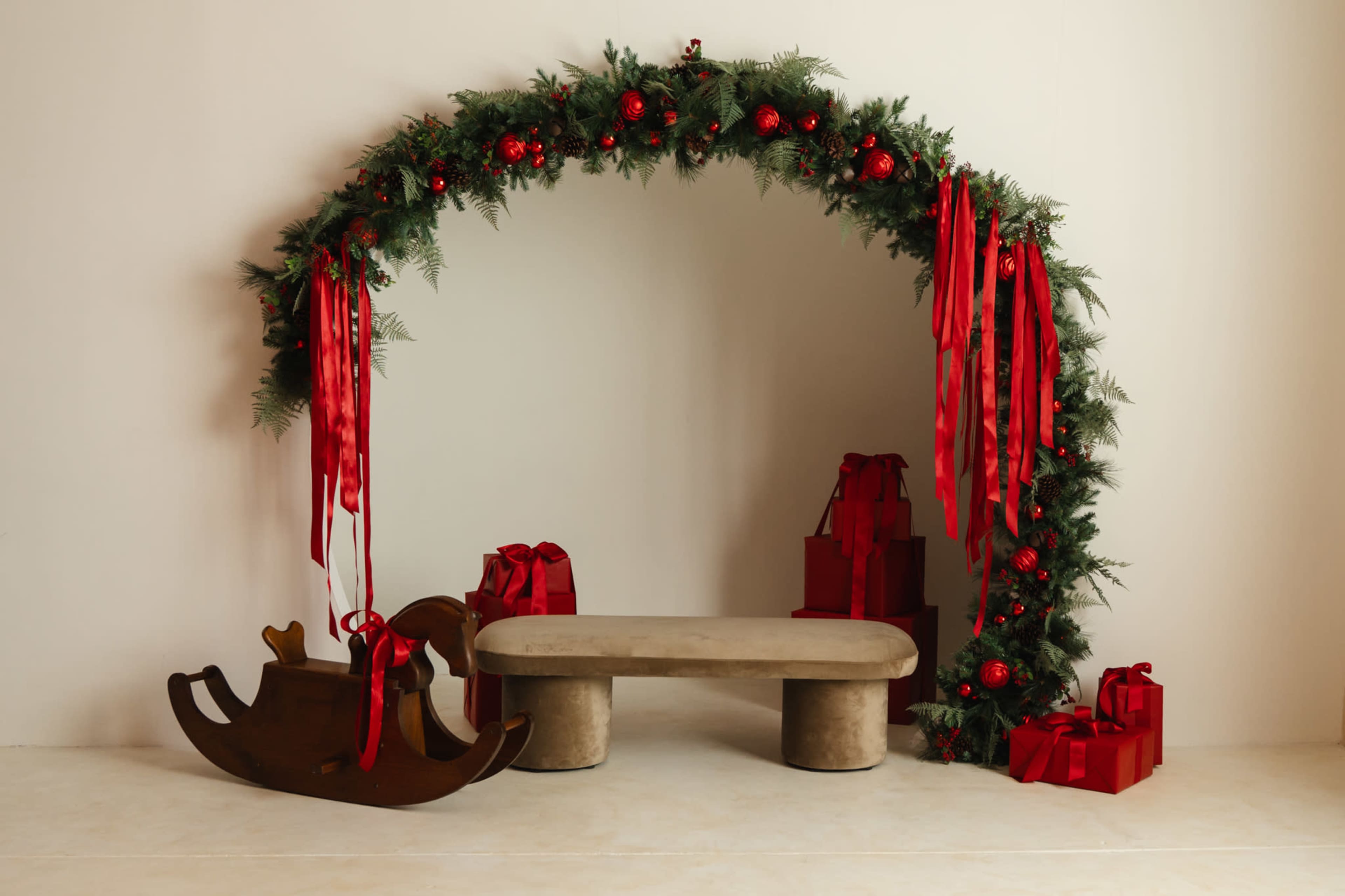 A festive arch made of greenery and red ornaments frames a simple stone bench, accompanied by a wooden rocking horse and neatly wrapped red gift boxes.