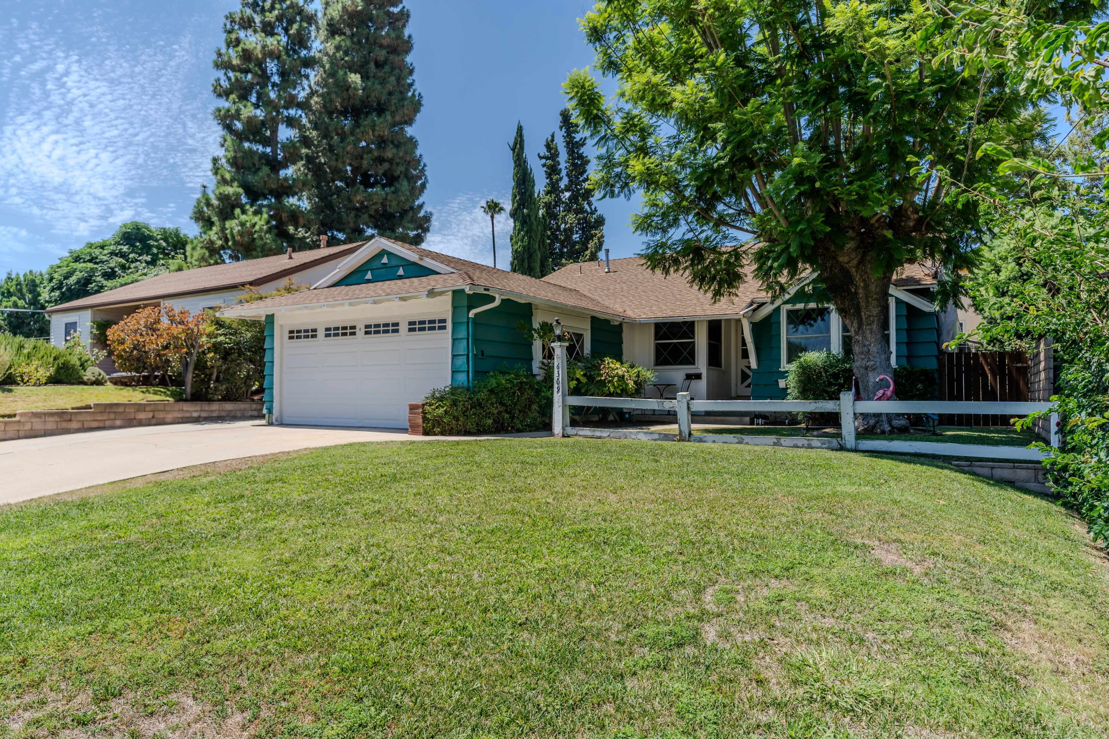 A single-story blue house with a garage and a neatly landscaped front yard is visible, surrounded by tall trees and shrubs.