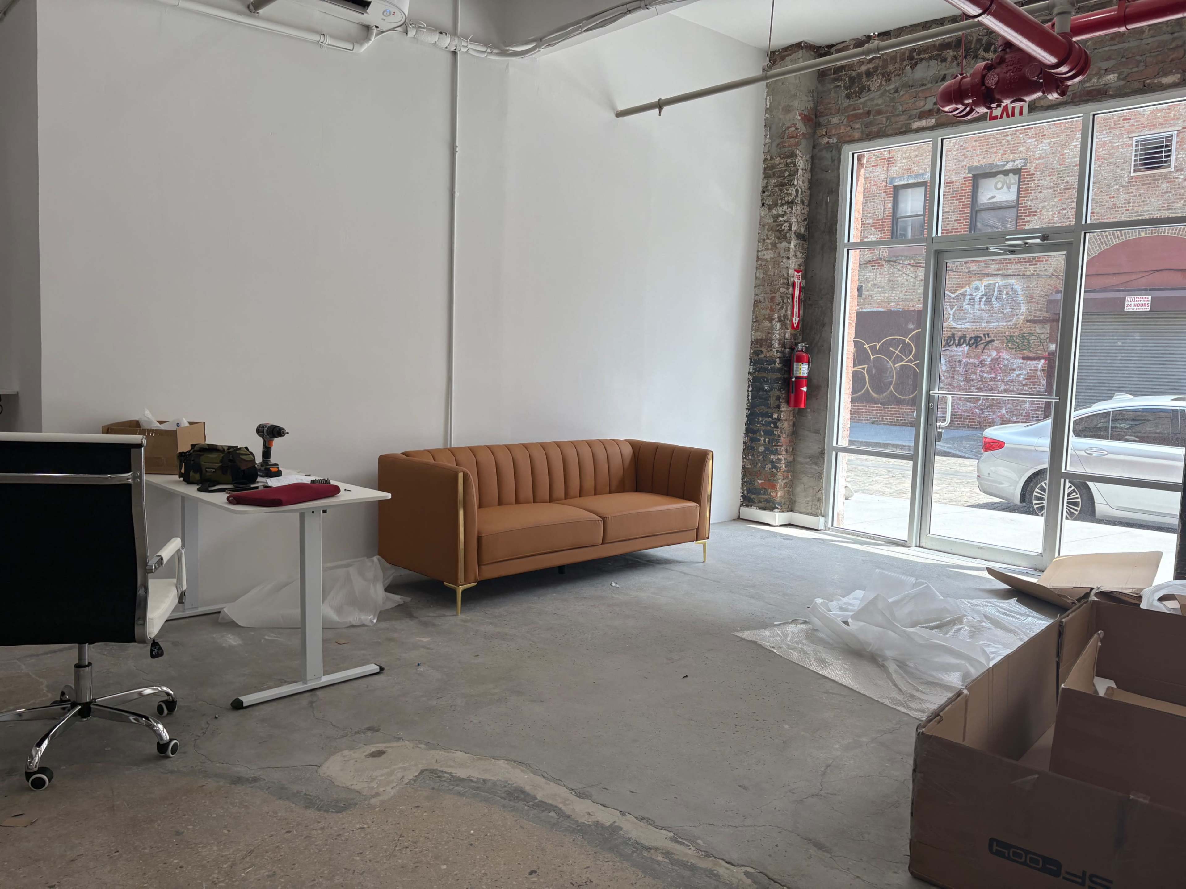 The image shows an empty room featuring a brown sofa, a white desk with a chair, and various packing materials scattered on the floor.