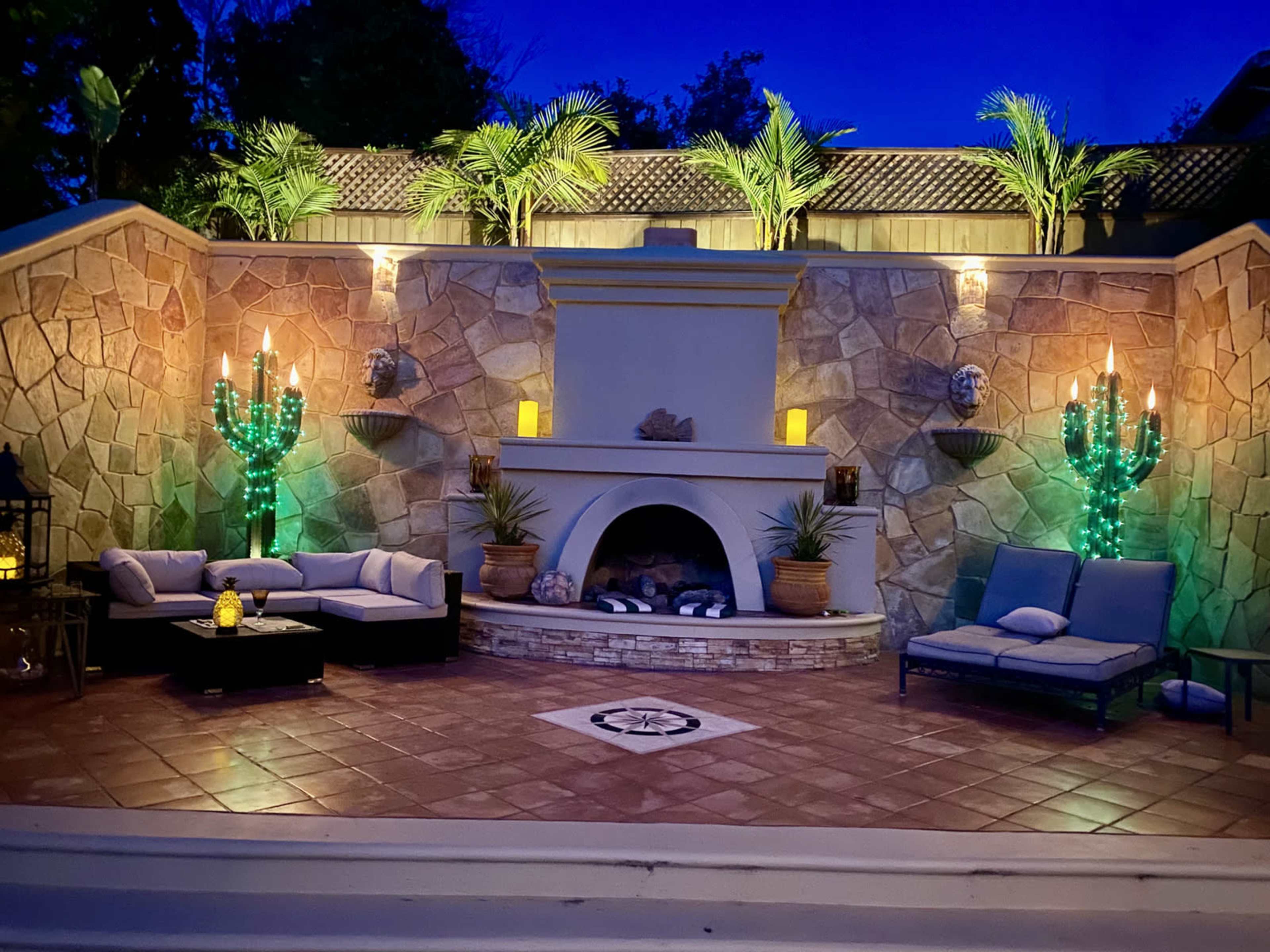 A well-lit outdoor seating area featuring two glowing cactus sculptures, a stone fireplace, and comfortable seating against a stone wall backdrop.