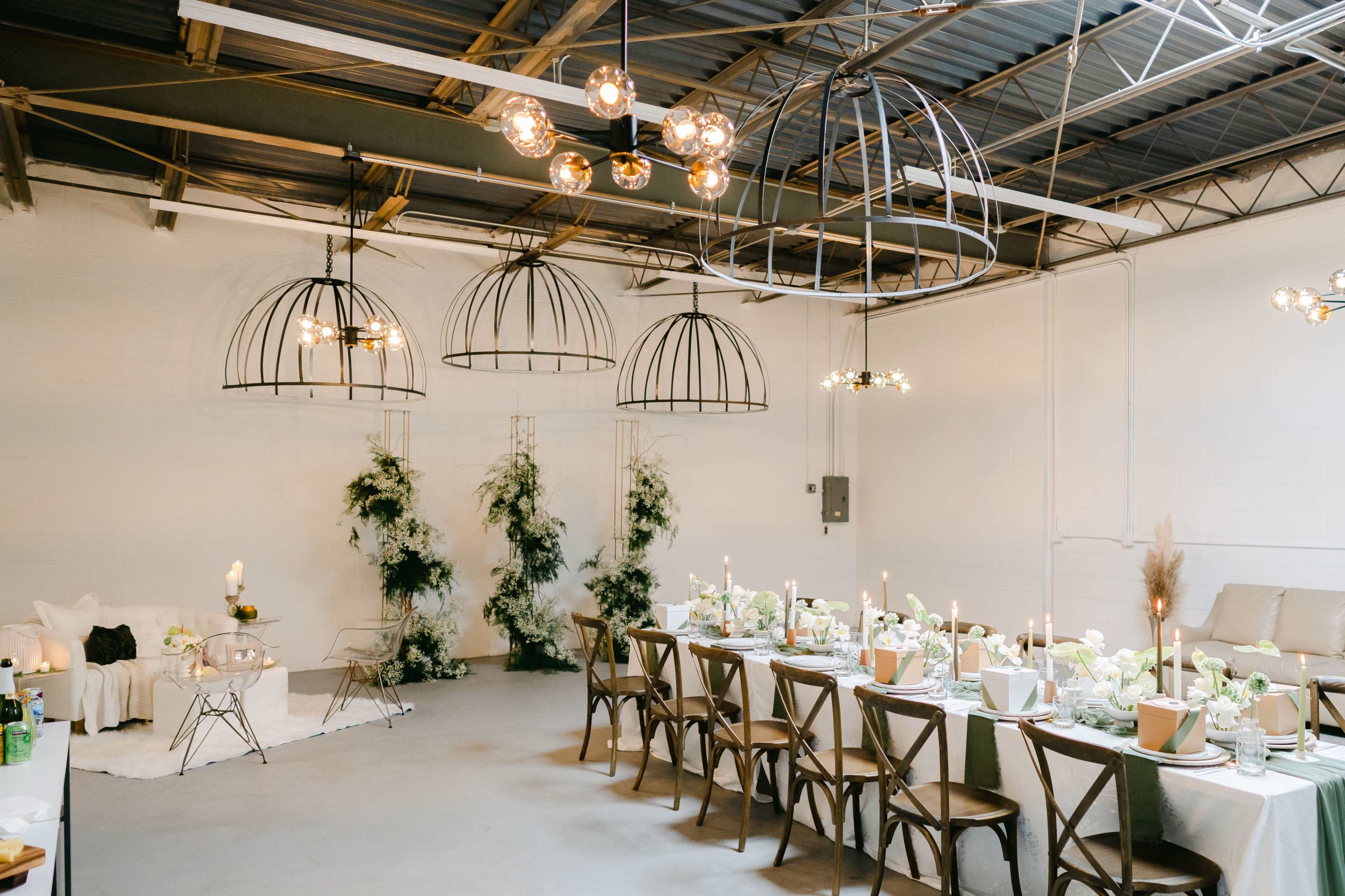 A dining area features a long table set with floral arrangements and candles, surrounded by industrial-style light fixtures and greenery accents.