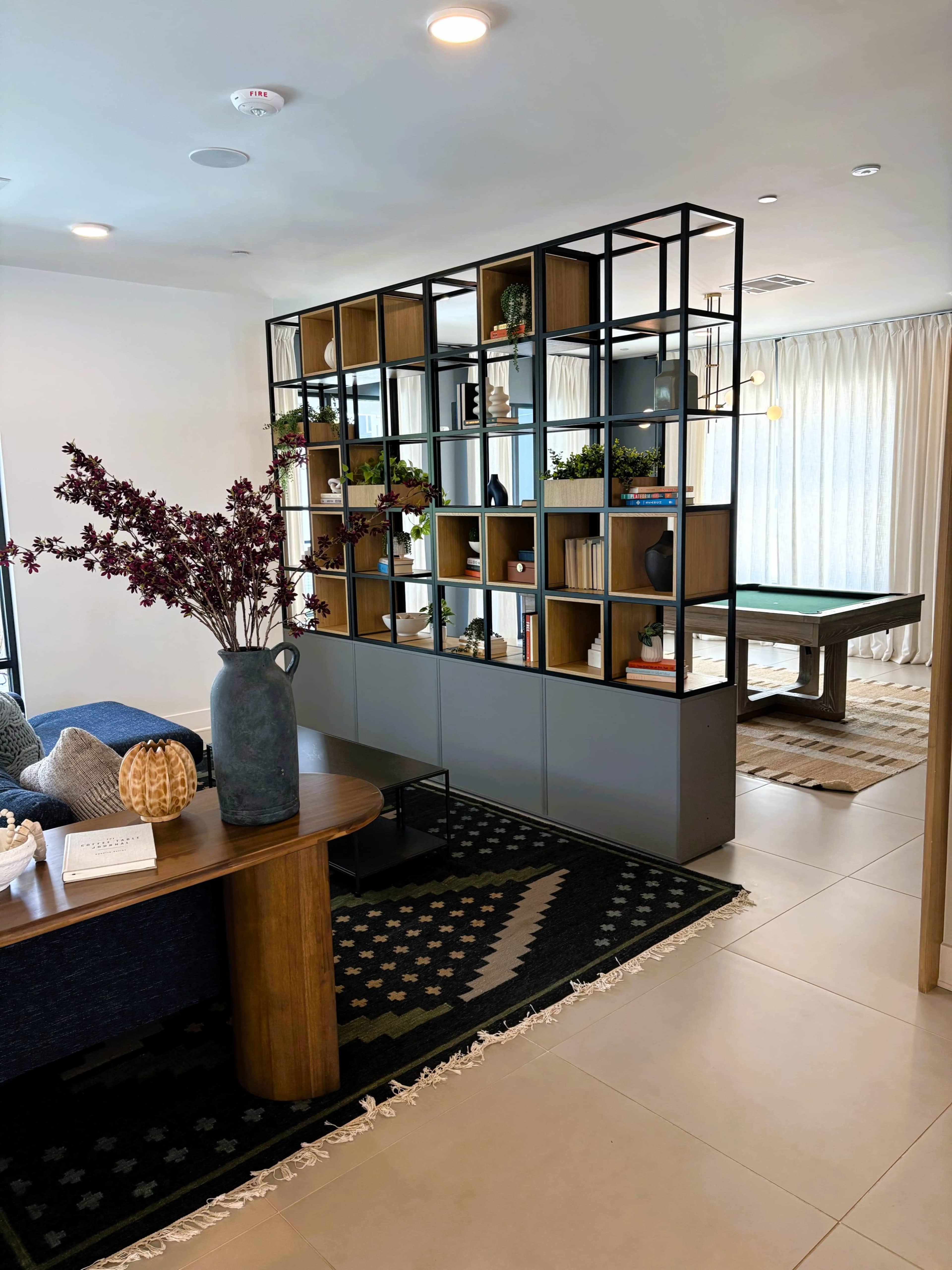 The image shows a modern interior space featuring a bookshelf divider, a wooden table with decorative items, and a pool table in the background.