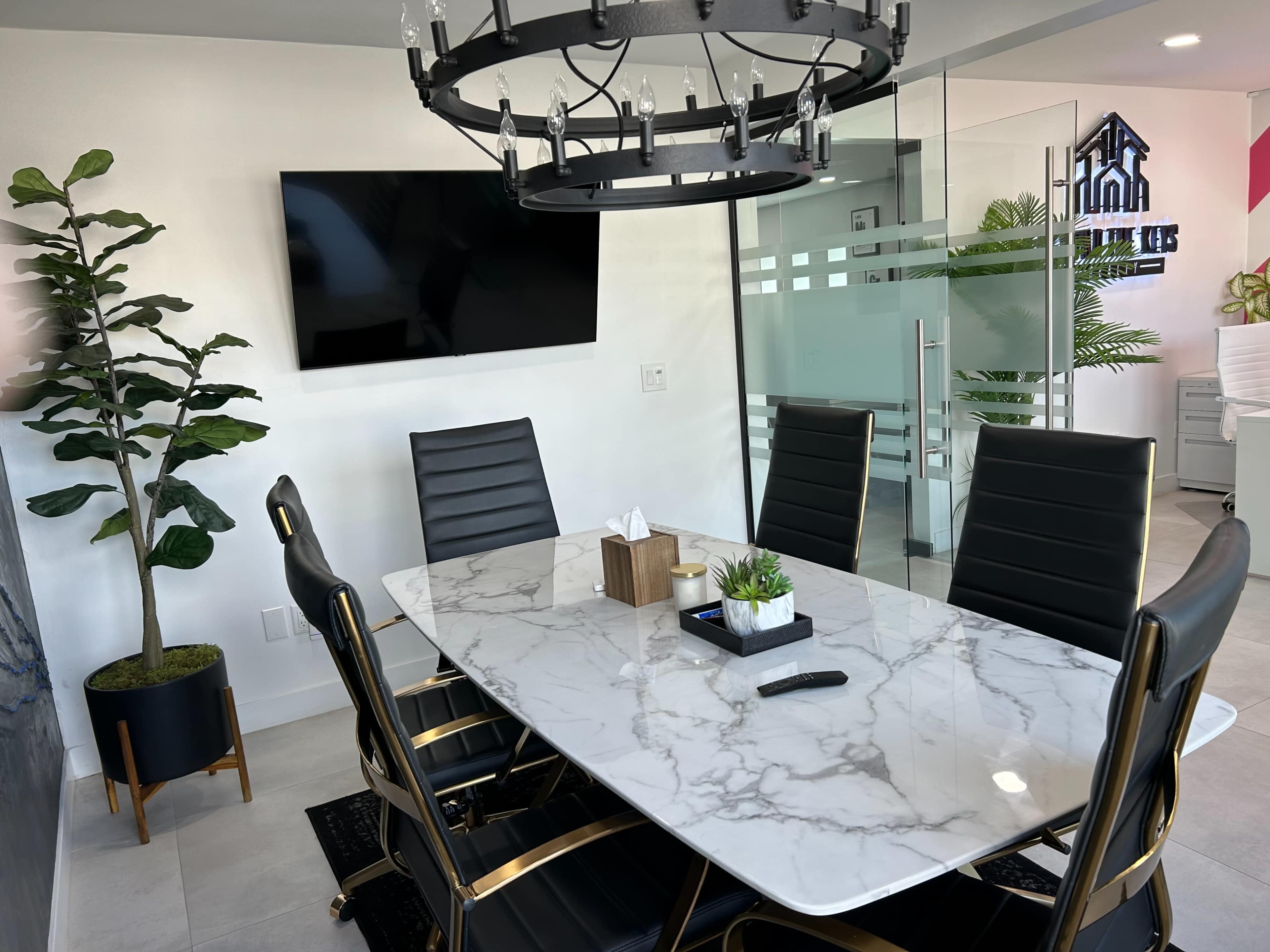 A modern meeting room featuring a marble dining table surrounded by black chairs, a wall-mounted television, and a decorative plant in a pot.