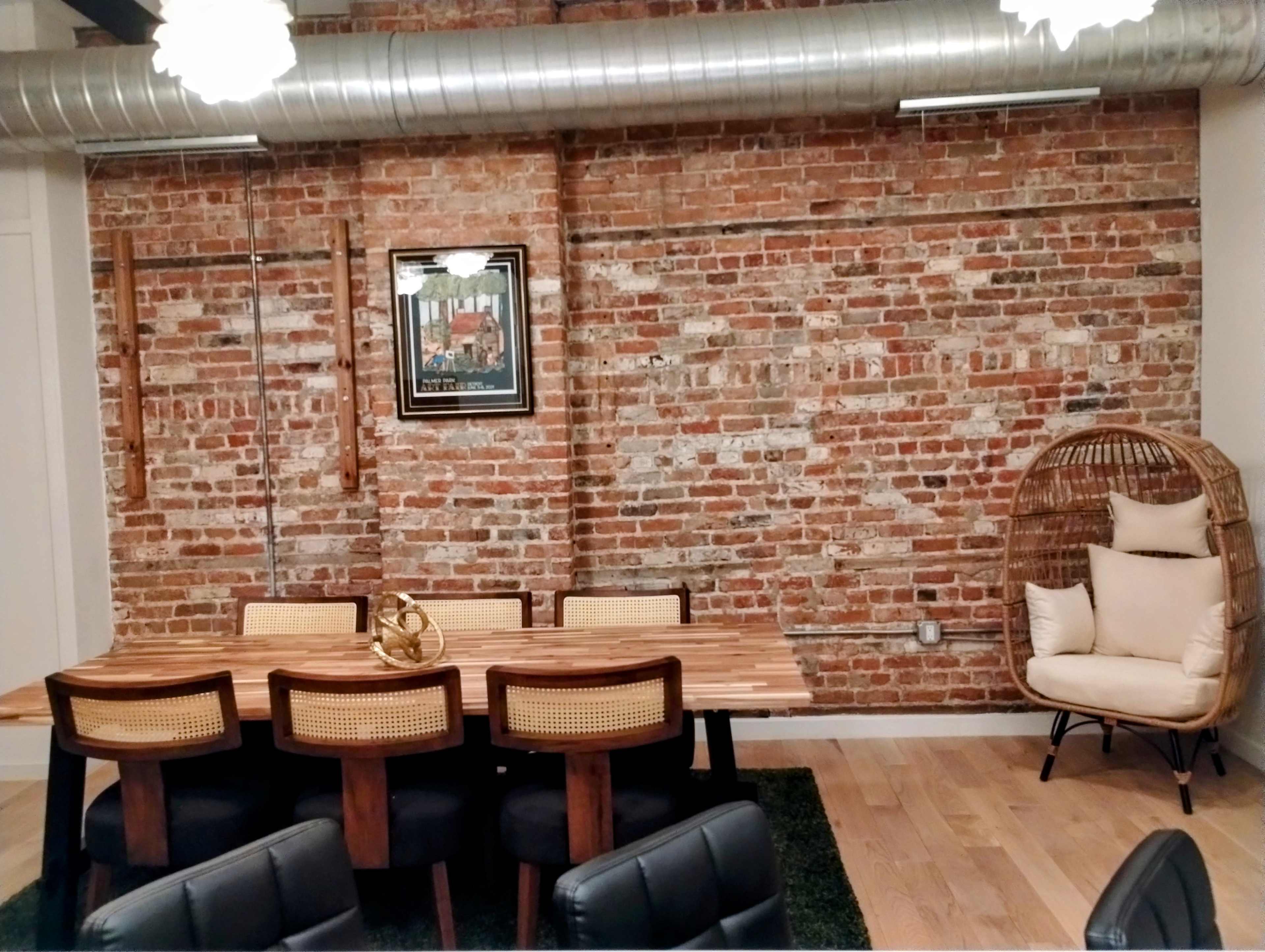 A dining area with a wooden table and chairs against a backdrop of exposed brick walls, featuring a decorative framed painting and a wicker chair in the corner.