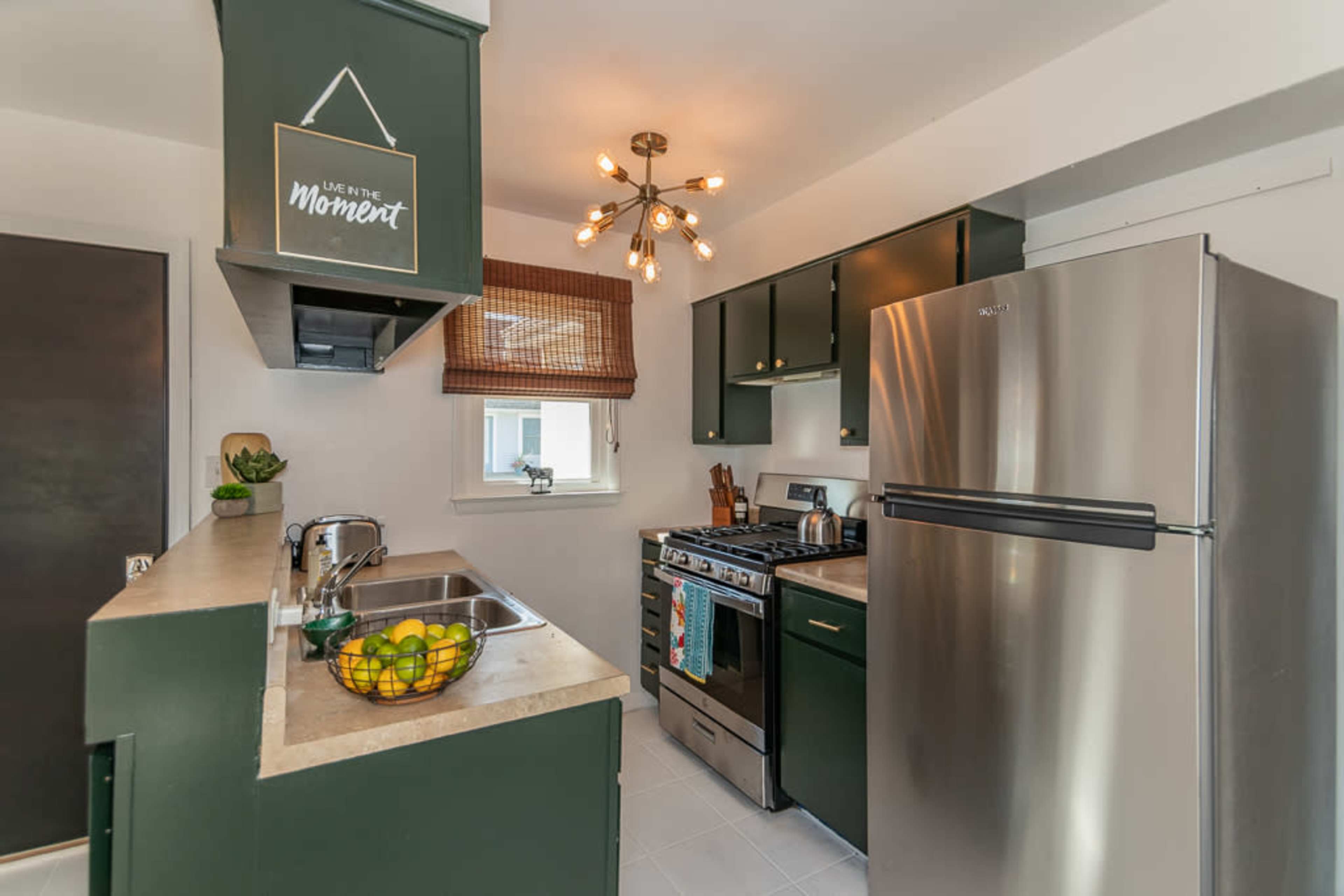The kitchen features dark green cabinetry, stainless steel appliances, and a central light fixture, with a small window and a bowl of lemons on the counter.