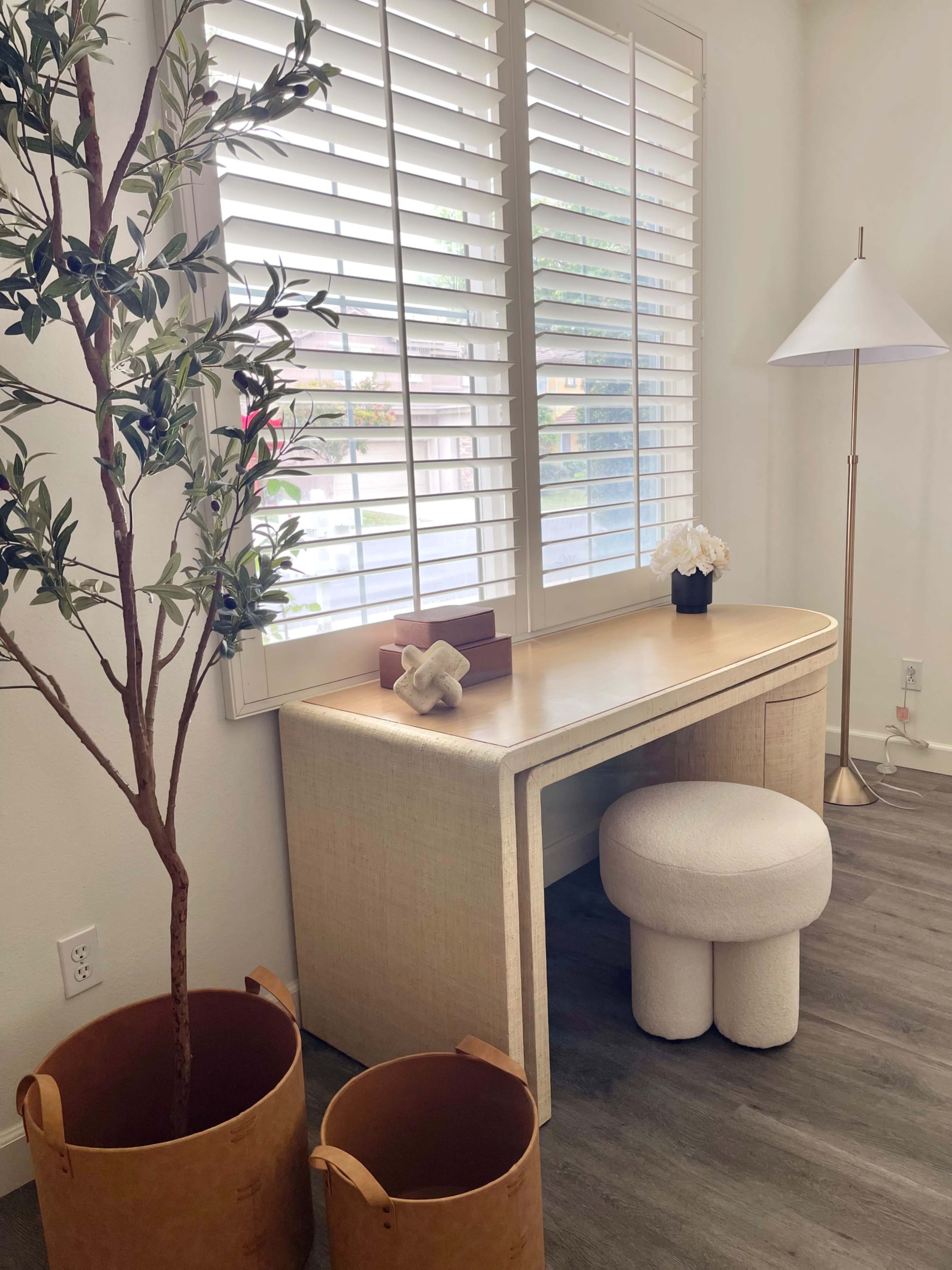 A minimalist workspace with a light-colored desk, a round stool, a decorative plant, and two brown containers on a wooden floor beside a window with plantation shutters.
