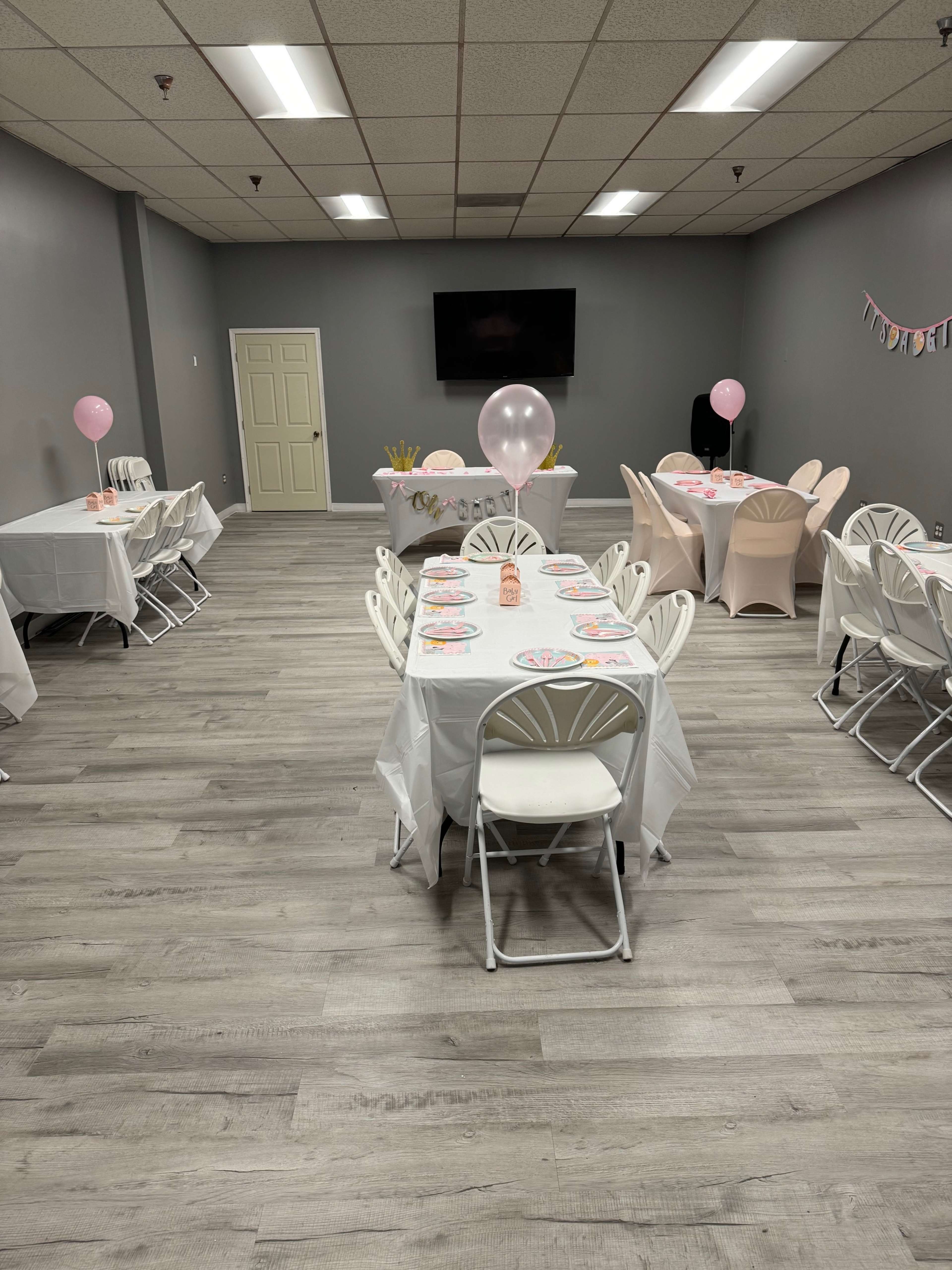 A room set up for a party, featuring several tables with white tablecloths and pink decorations, along with balloons and a television on the wall.
