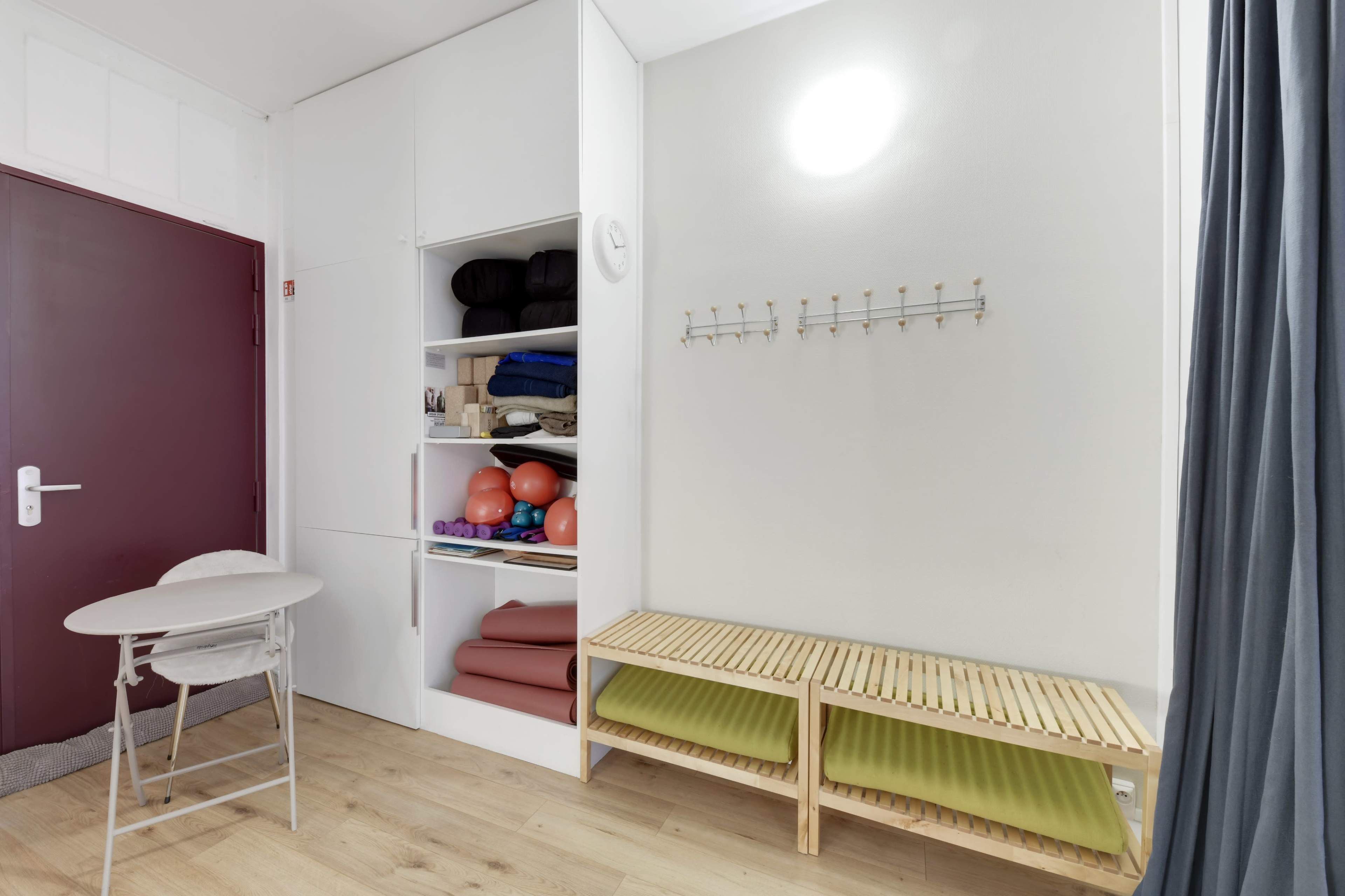 The room features a compact storage area with shelves containing exercise mats and equipment, a small round table, and a wooden bench along the wall.