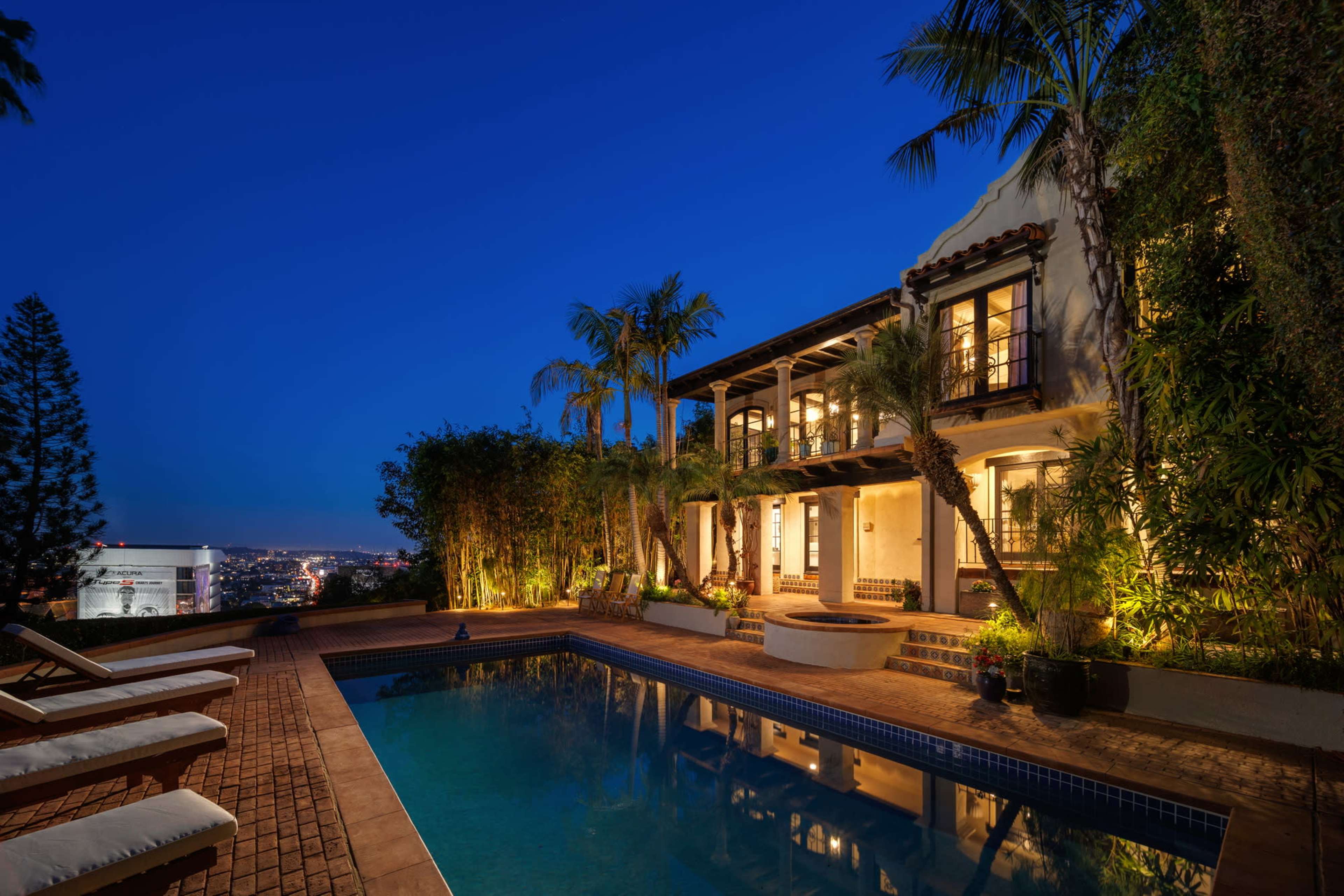 A two-story villa with a pool and landscaped surroundings is illuminated at night, showcasing a view of the city in the background.