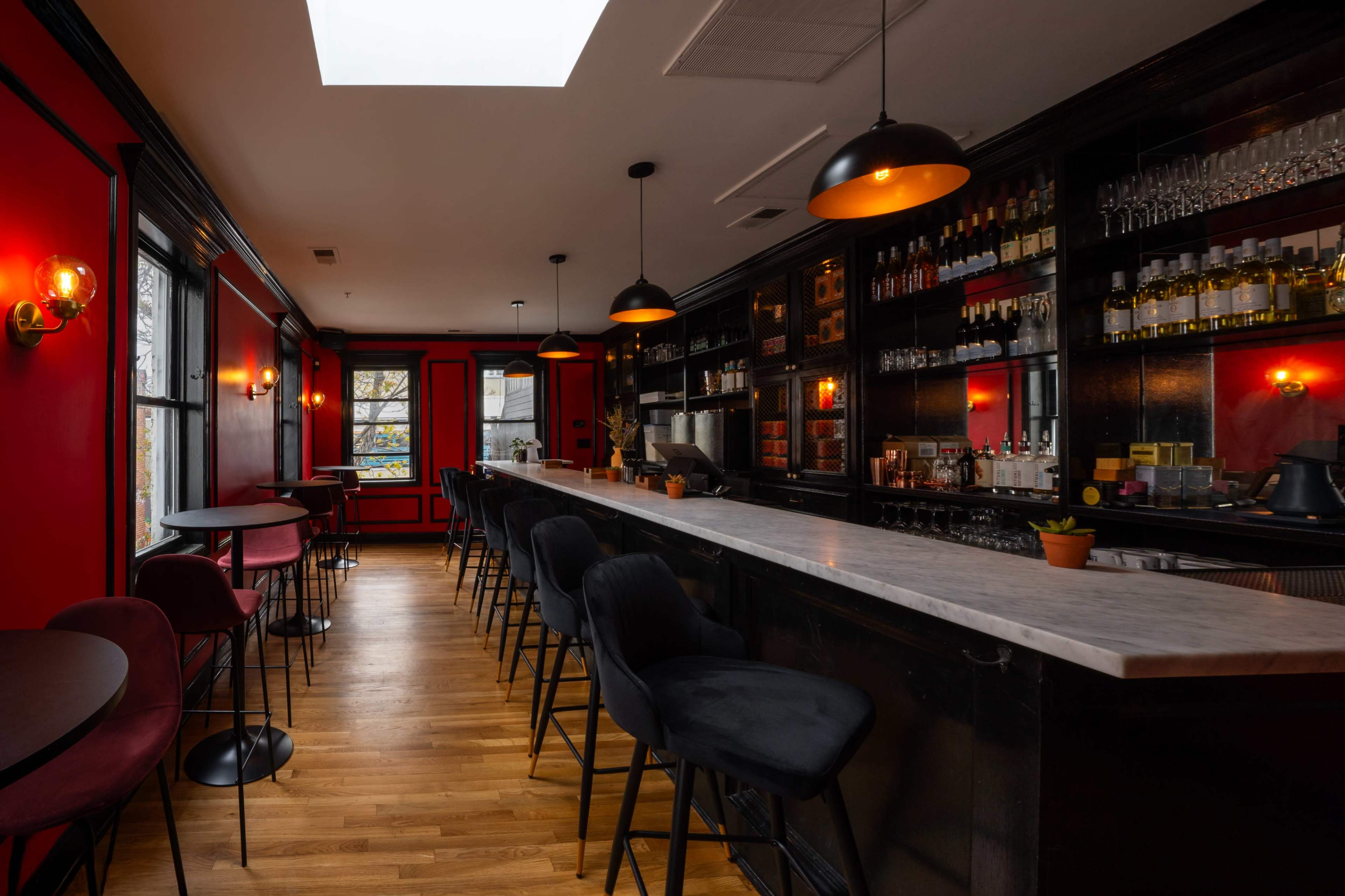 The image shows a stylish bar with a marble countertop, black shelving filled with liquor bottles, and red walls illuminated by hanging lights.