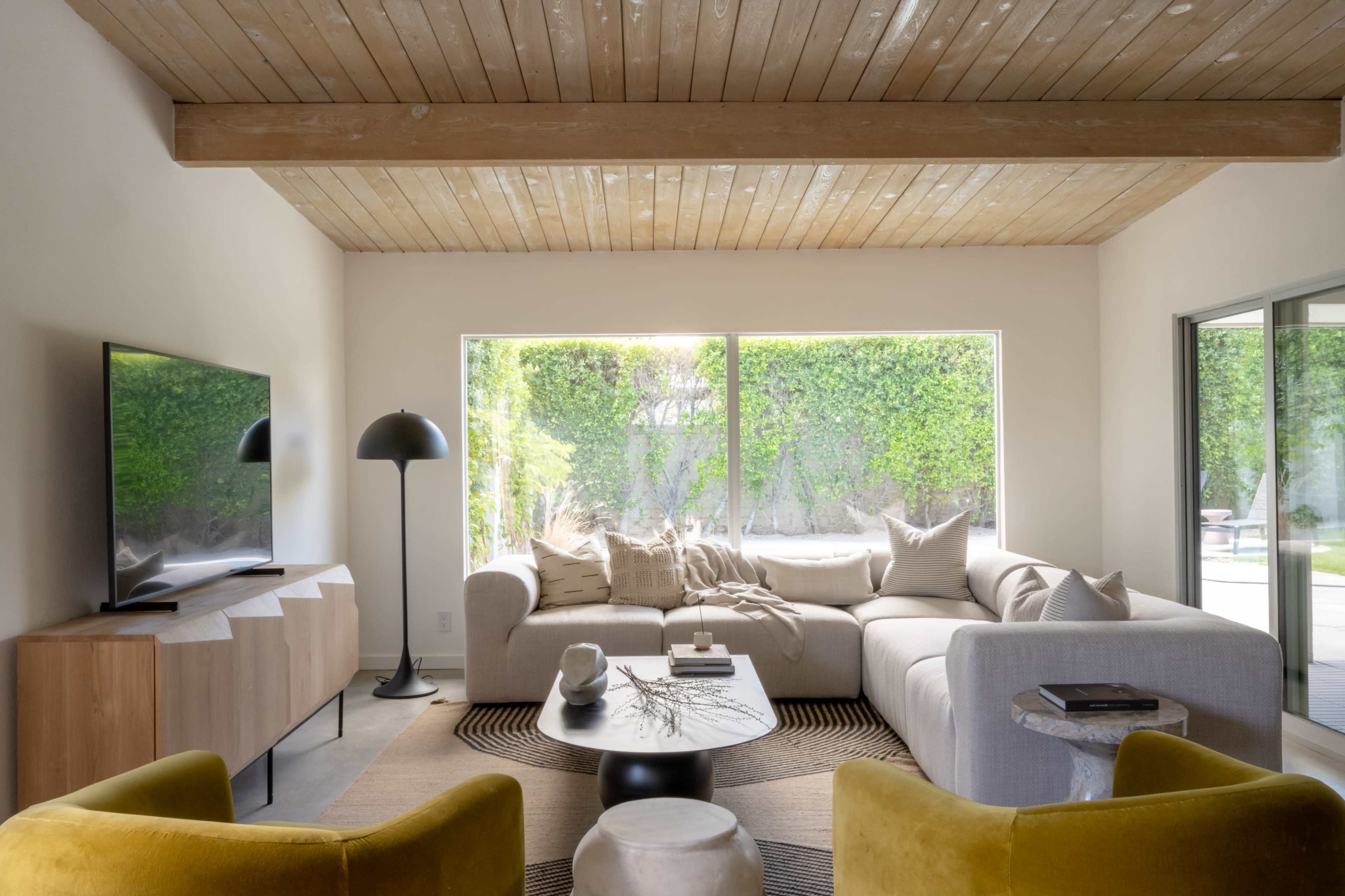 A modern living room features a light-colored sectional sofa, a round coffee table, and a large window overlooking a green outdoor space.
