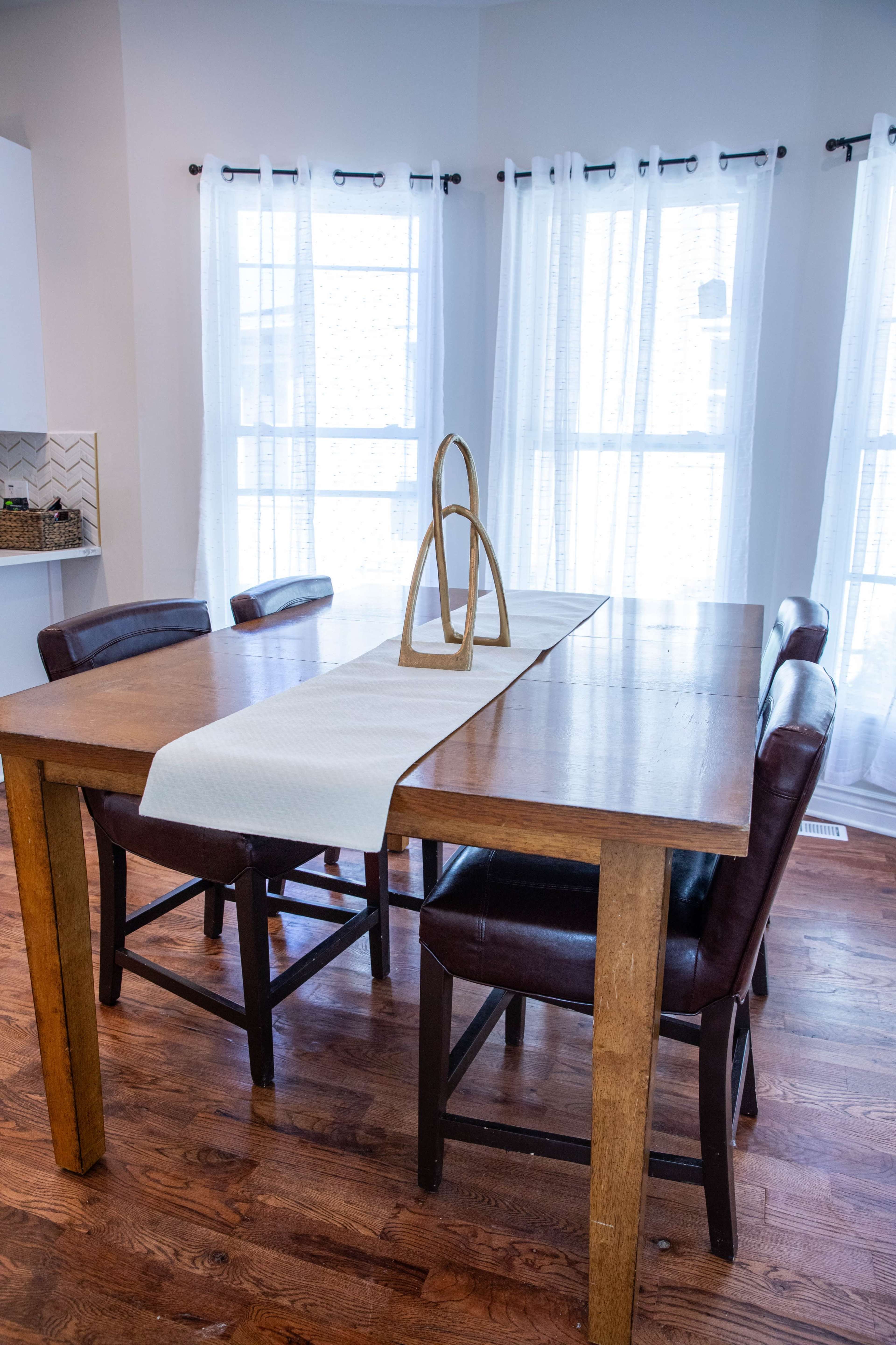 A wooden dining table with a light-colored runner is surrounded by dark leather chairs in a well-lit room with large windows.