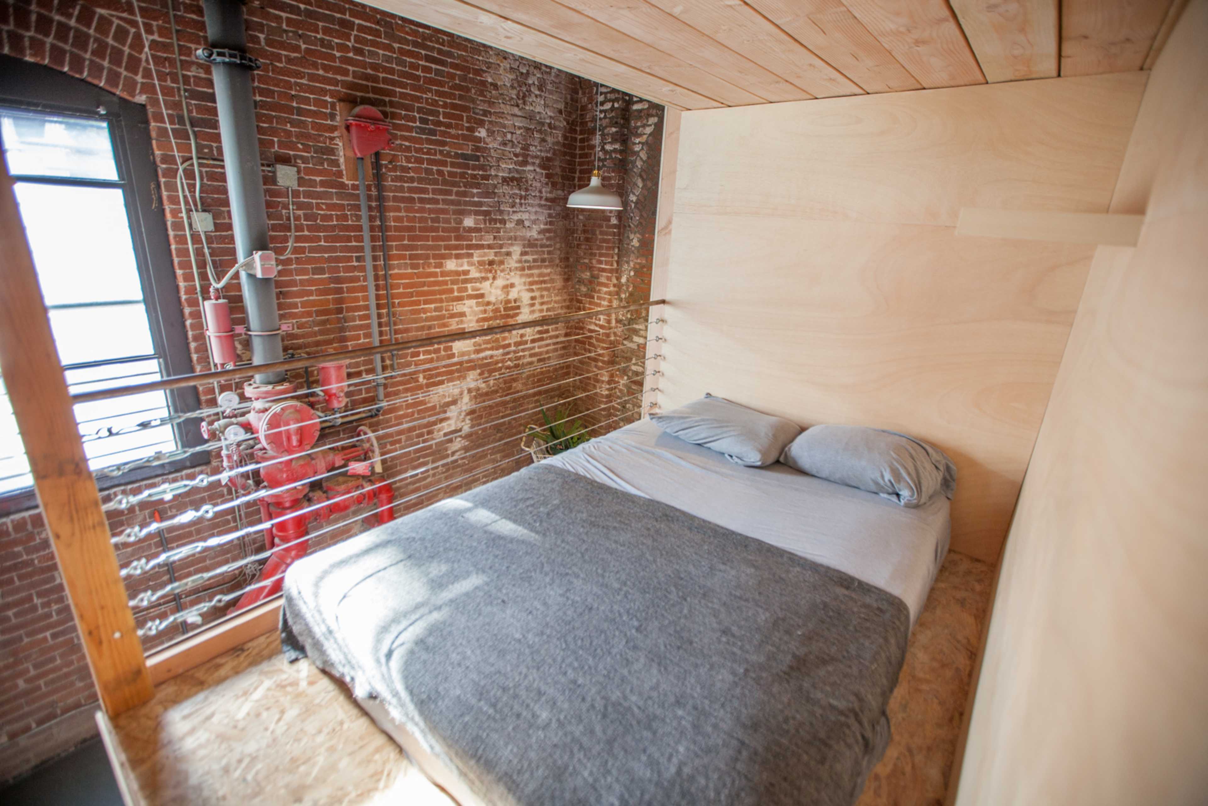 A minimalist loft bedroom with a wooden frame, a bed with gray bedding, and exposed brick walls.