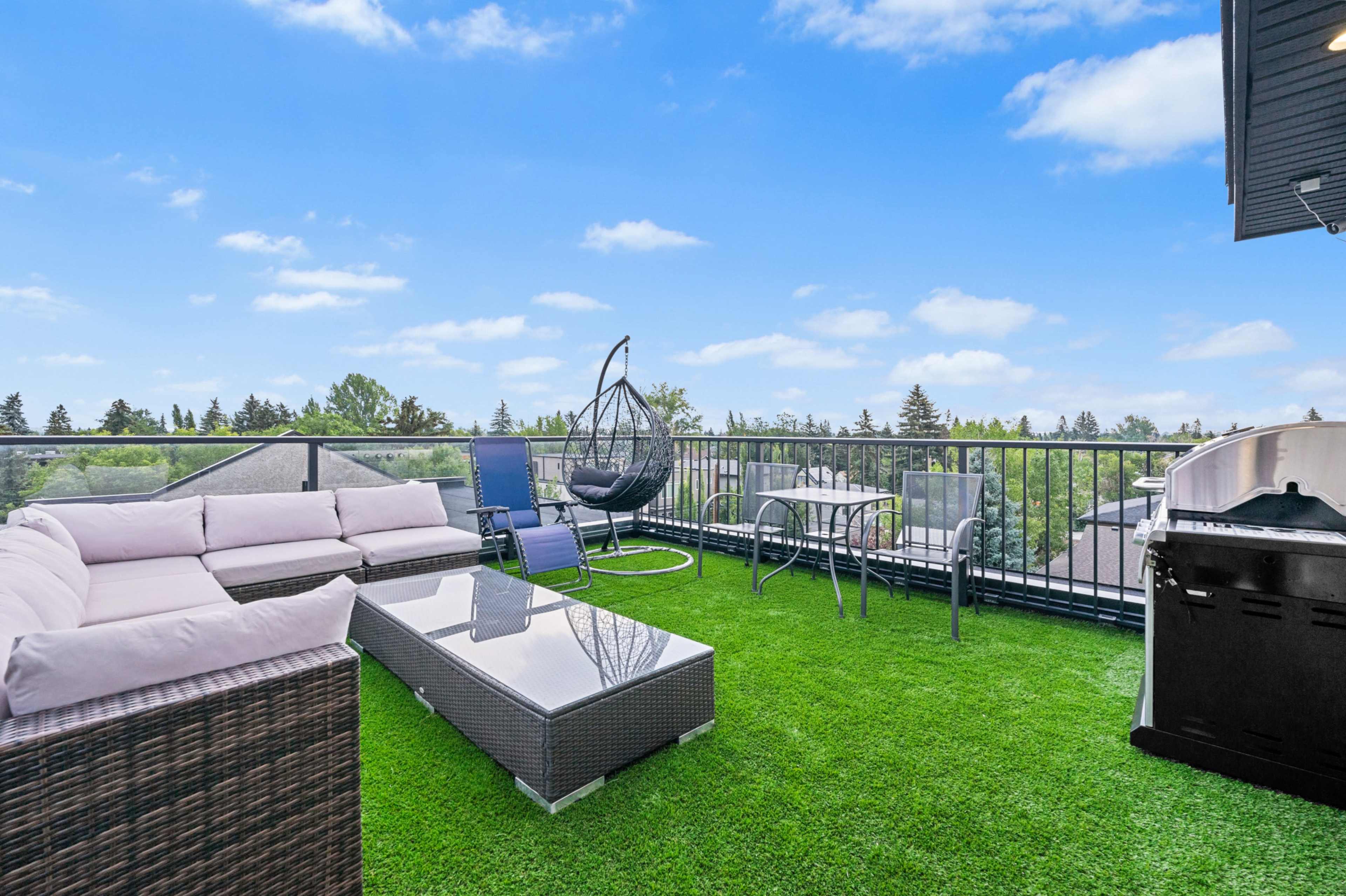 The image shows a rooftop terrace featuring a sectional sofa, a hanging chair, a glass coffee table, and a dining set on artificial grass.