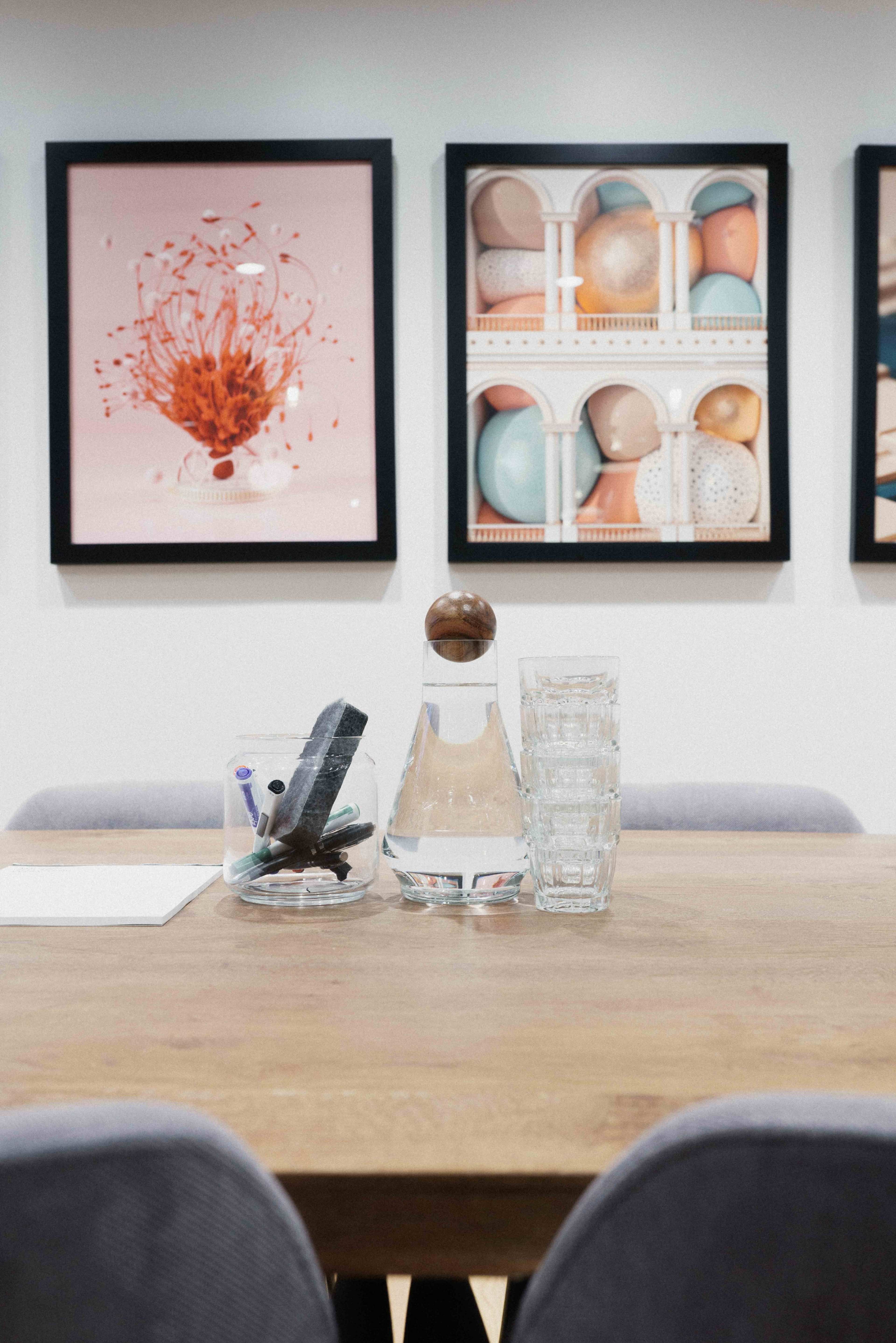 A wooden table with a glass carafe and stacked glasses is set in a room displaying framed artwork on the walls.