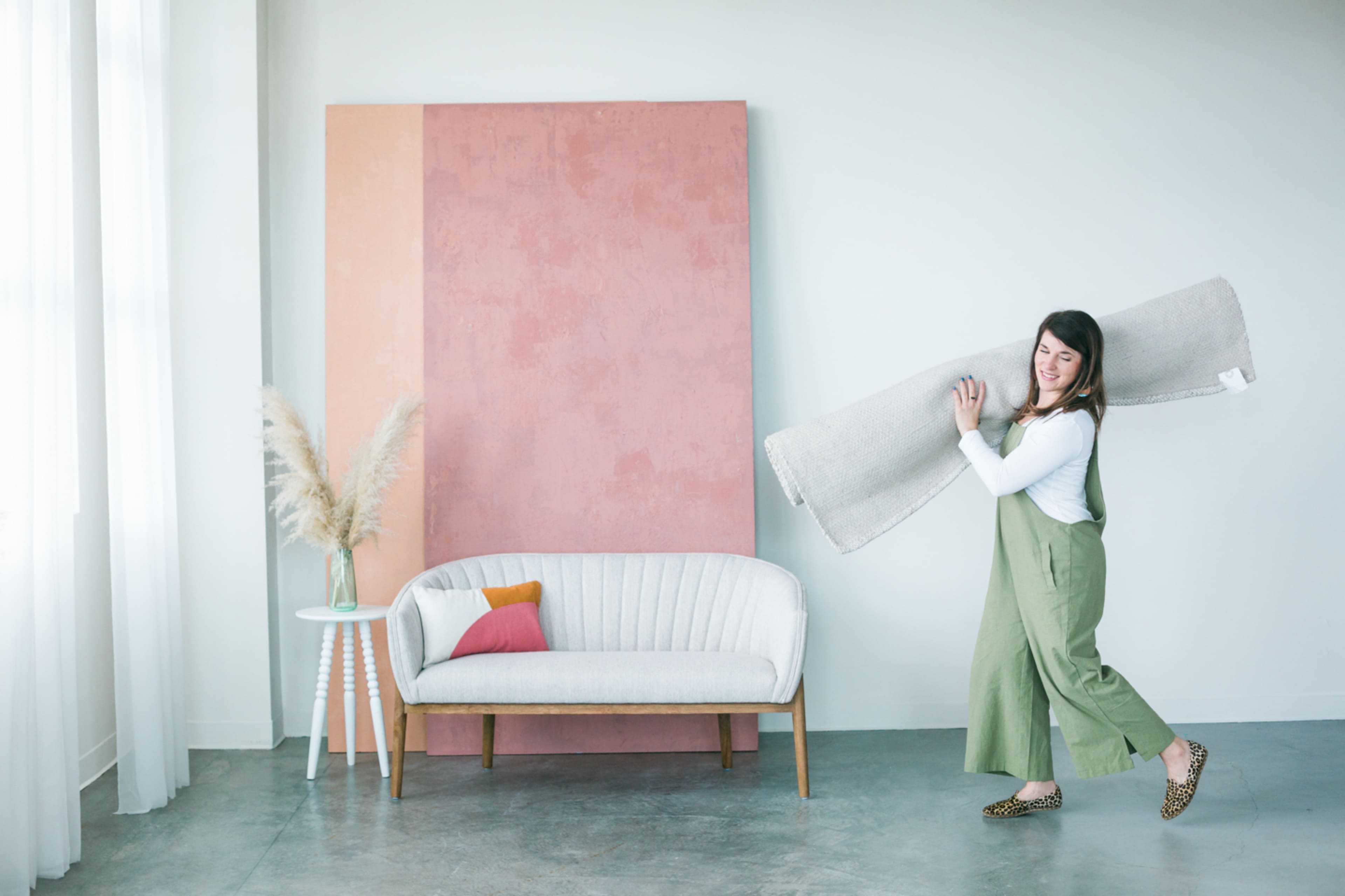 A woman in green overalls carries a large rolled rug while walking past a light-colored couch and a pink and beige wall.