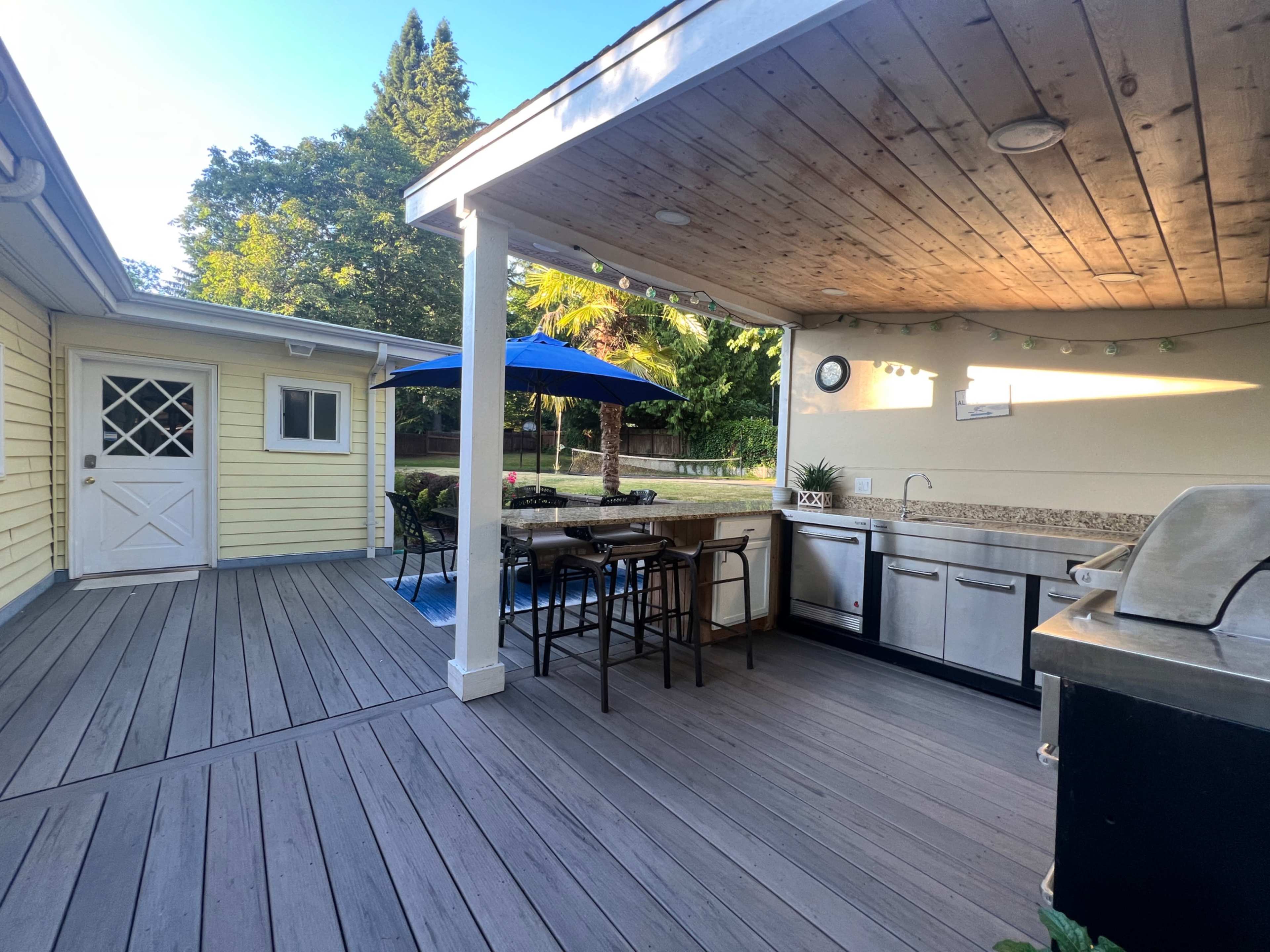 The image shows an outdoor kitchen area with a grill and seating on a wooden deck, adjacent to a yellow house and a shaded dining table under a blue umbrella.