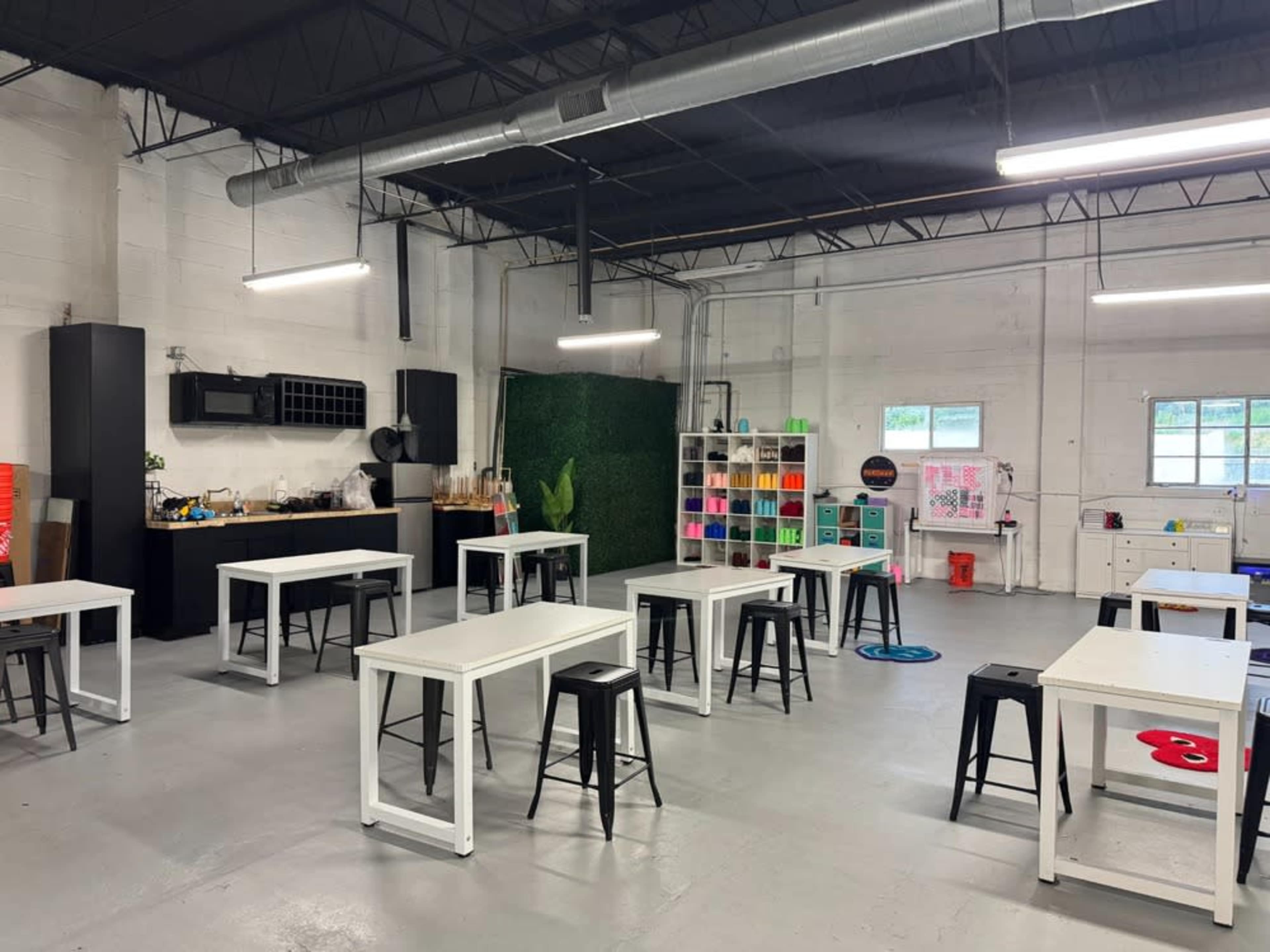 The image shows a spacious workshop area with several tables and chairs arranged in a clean, well-lit environment, accompanied by a kitchenette and shelves filled with colorful supplies.