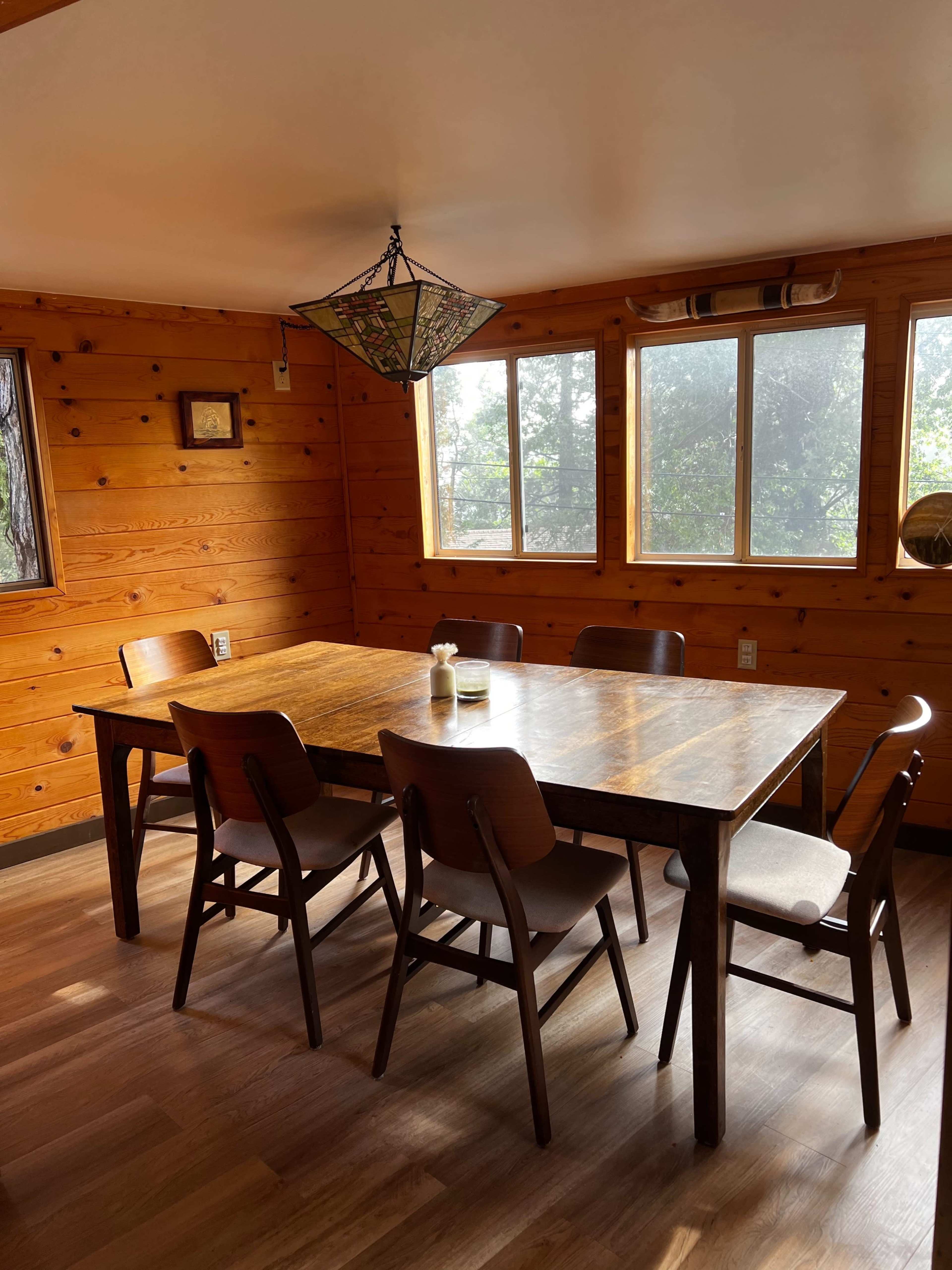 A wooden dining table with six chairs is set in a room with wooden walls and large windows letting in natural light.