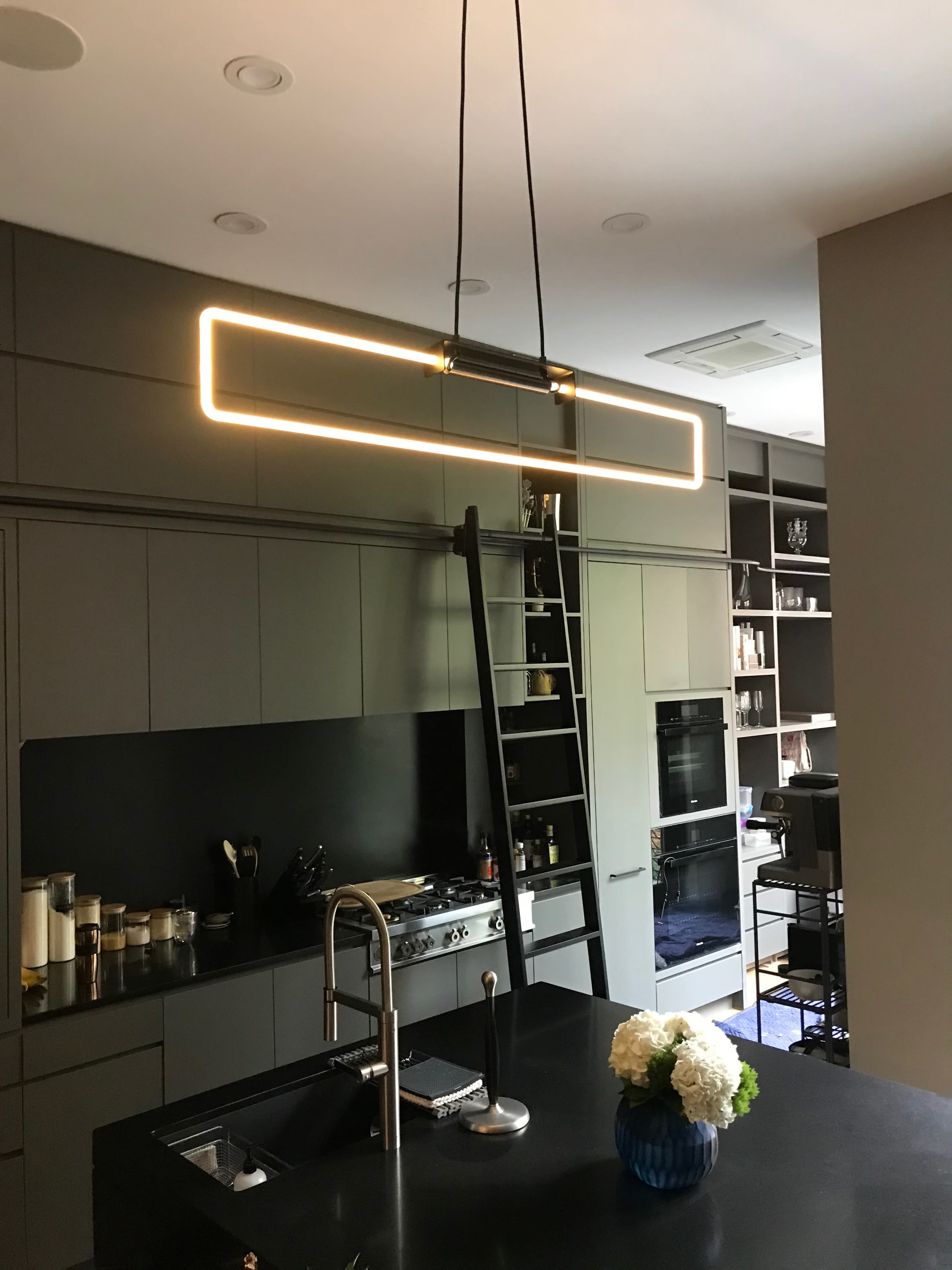 A modern kitchen features a central island with a black countertop and a rectangular pendant light hanging above, surrounded by sleek cabinetry and shelving units.