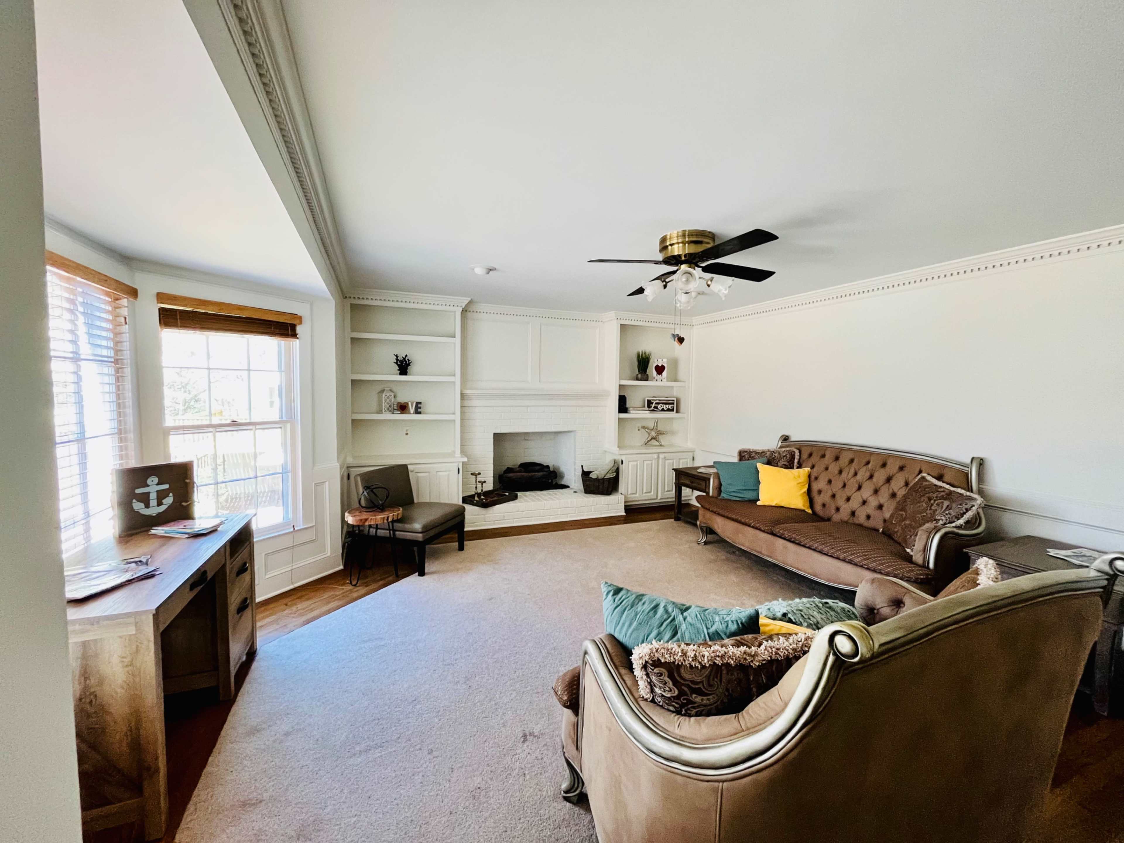 A living room featuring a brown sofa, two armchairs, a fireplace, built-in shelves, and large windows with wooden blinds.