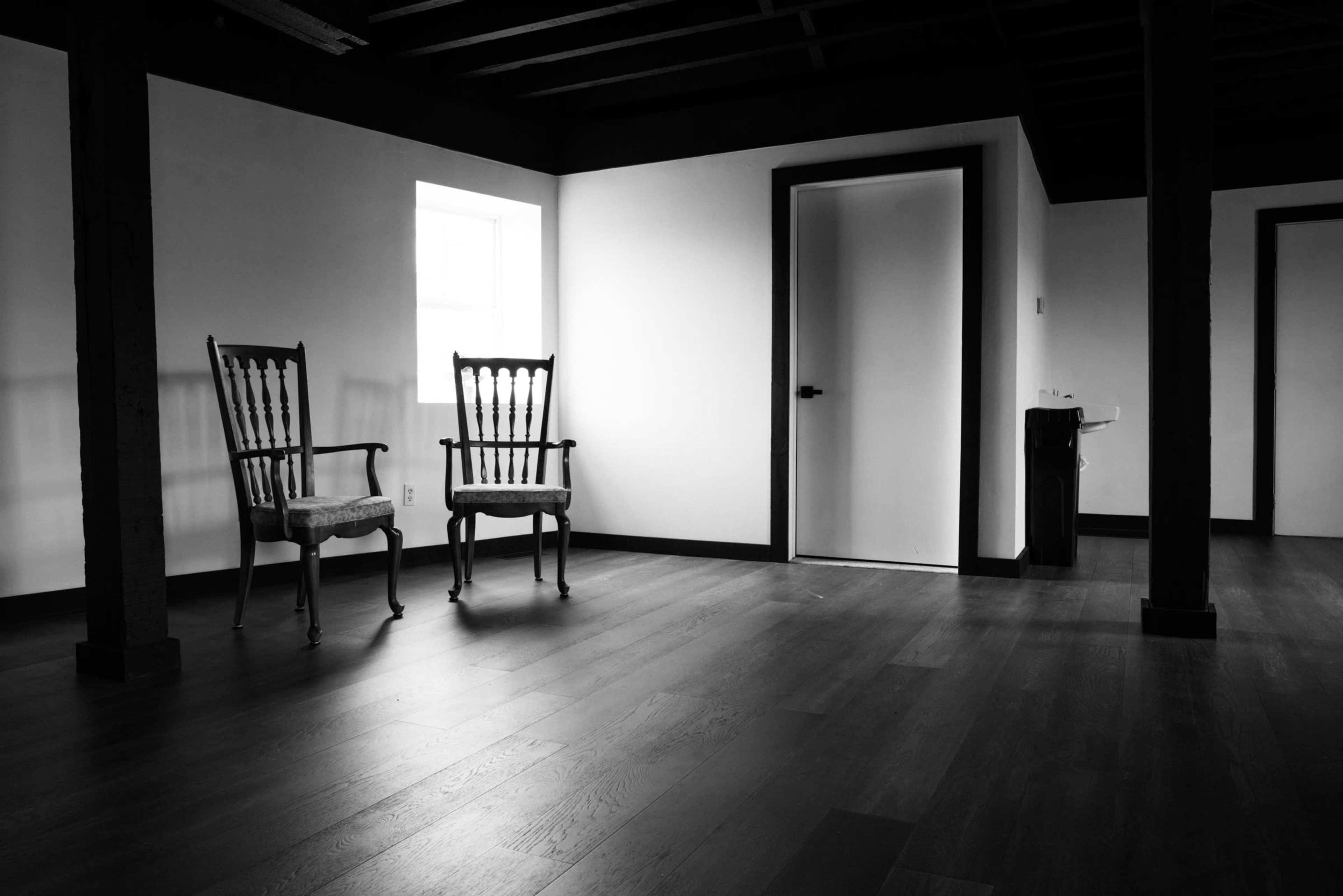 The image shows an empty room with two wooden chairs facing a door and a window, featuring dark wooden beams and a light-colored wall.