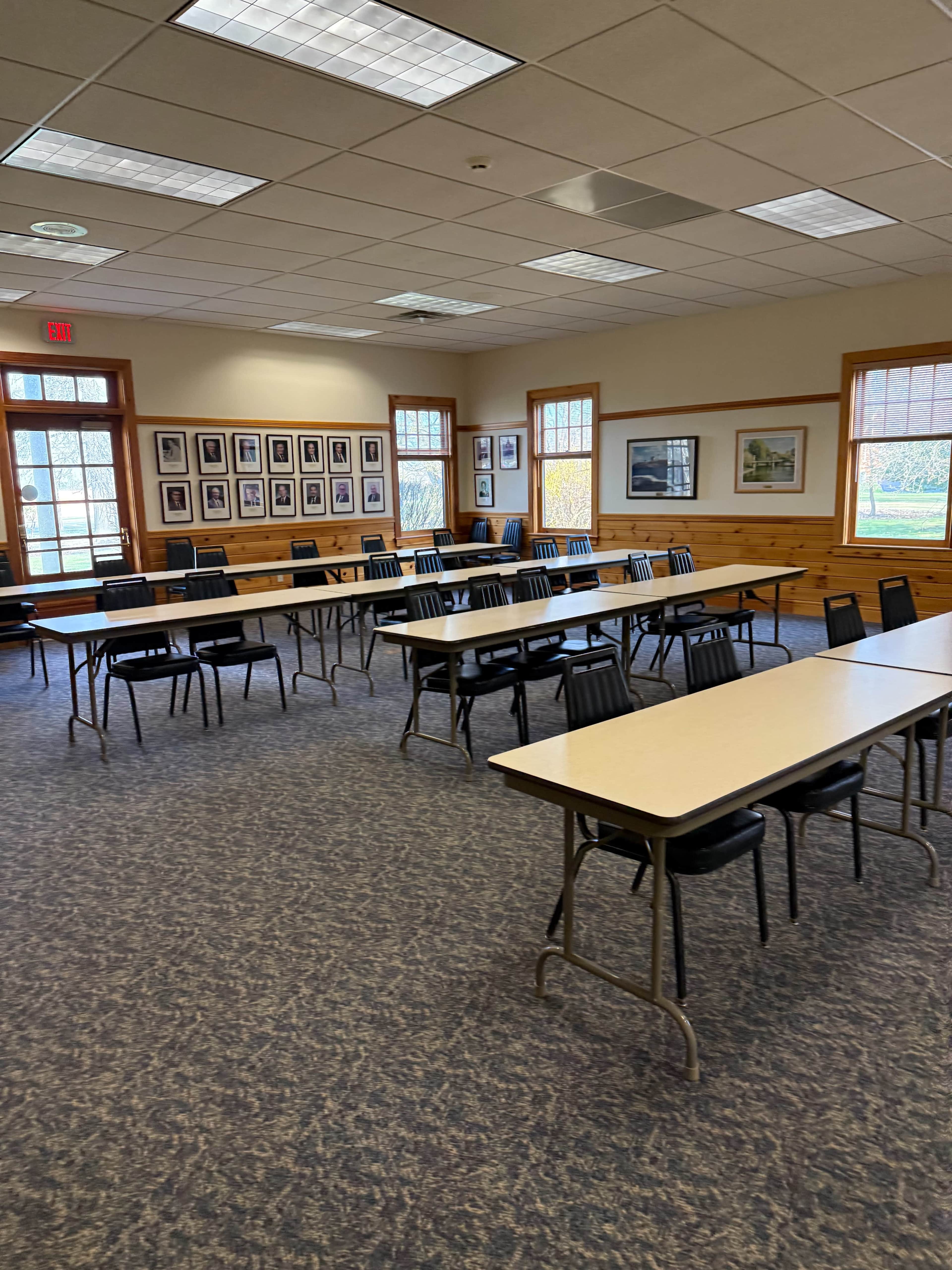 The image shows a room with several rows of tables and chairs arranged for a meeting or event, featuring windows with natural light and framed photographs on the walls.