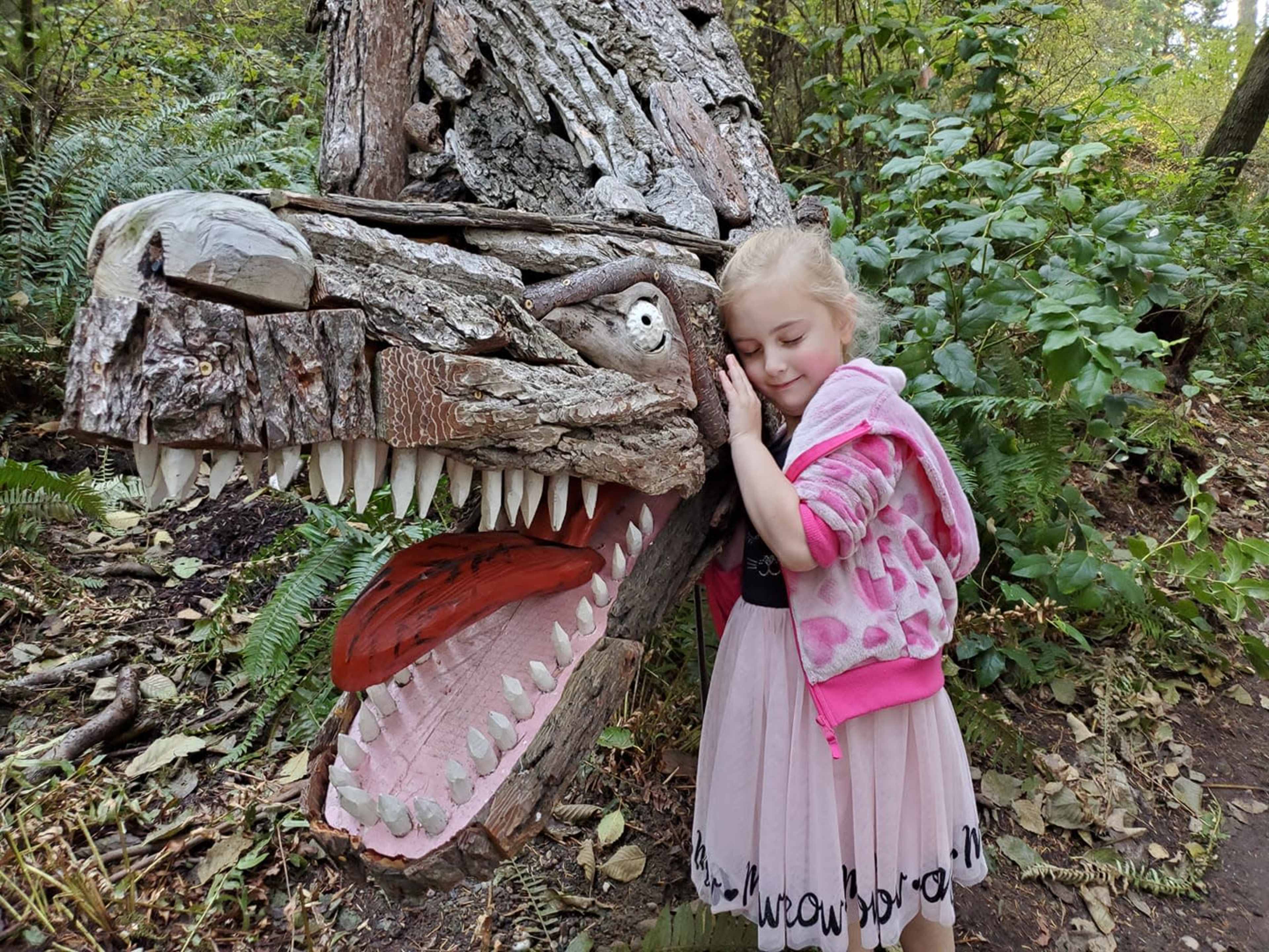 A child leans against a carved wooden dragon's head in a forested area.