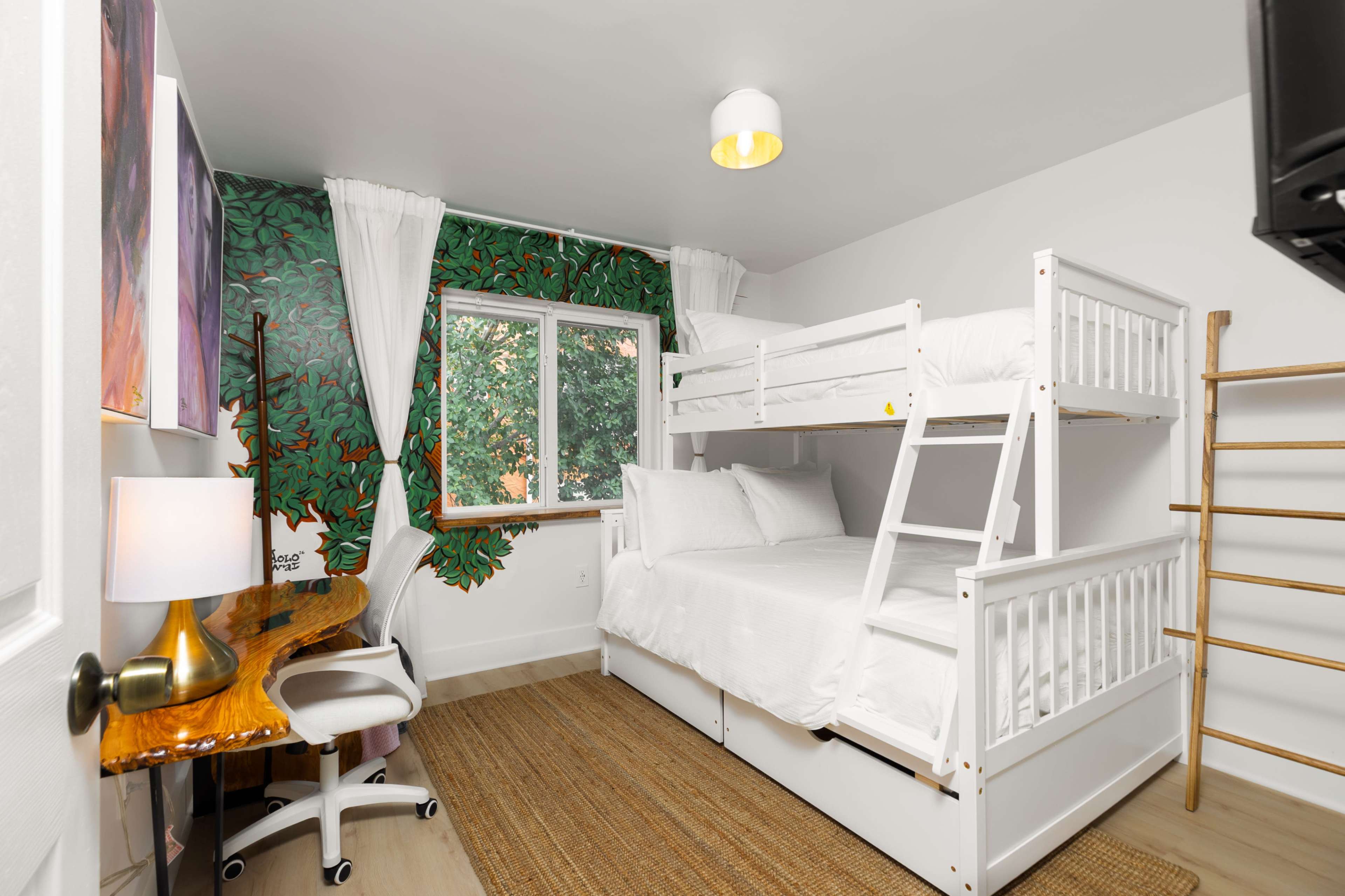 The room features a bunk bed against a wall decorated with a large green tree mural, a wooden desk, and a window with sheer white curtains.