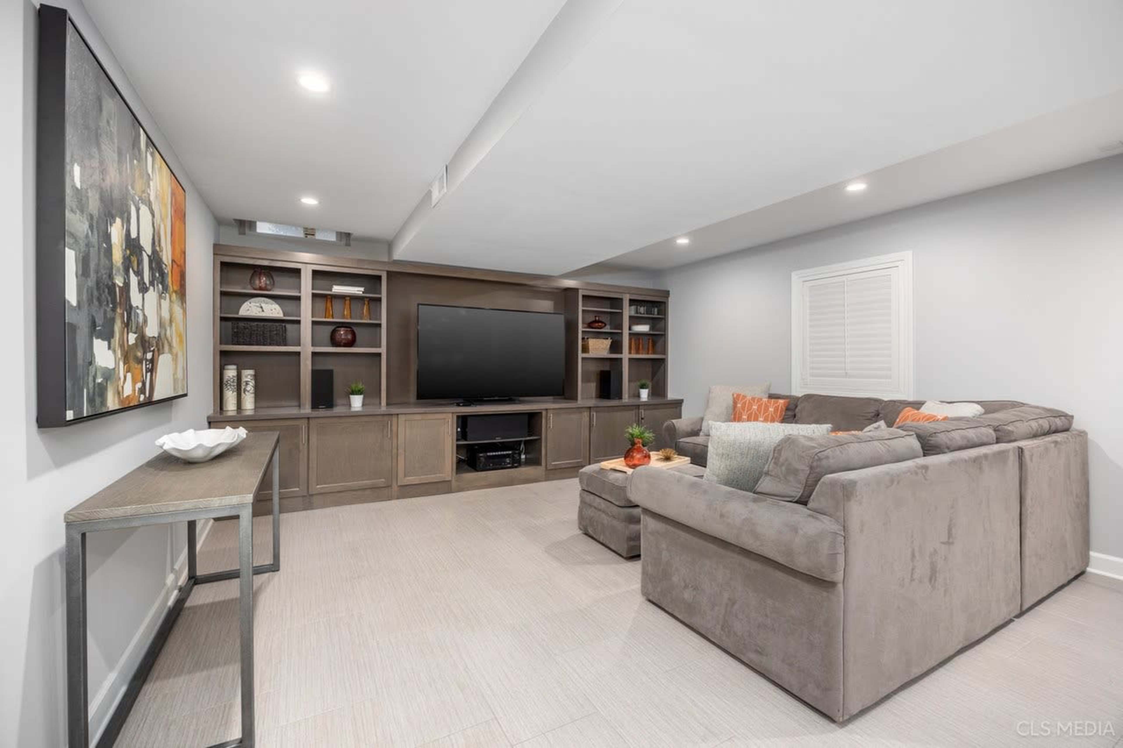 The image shows a well-lit basement living area featuring a large sectional sofa, a mounted television, and built-in shelving.