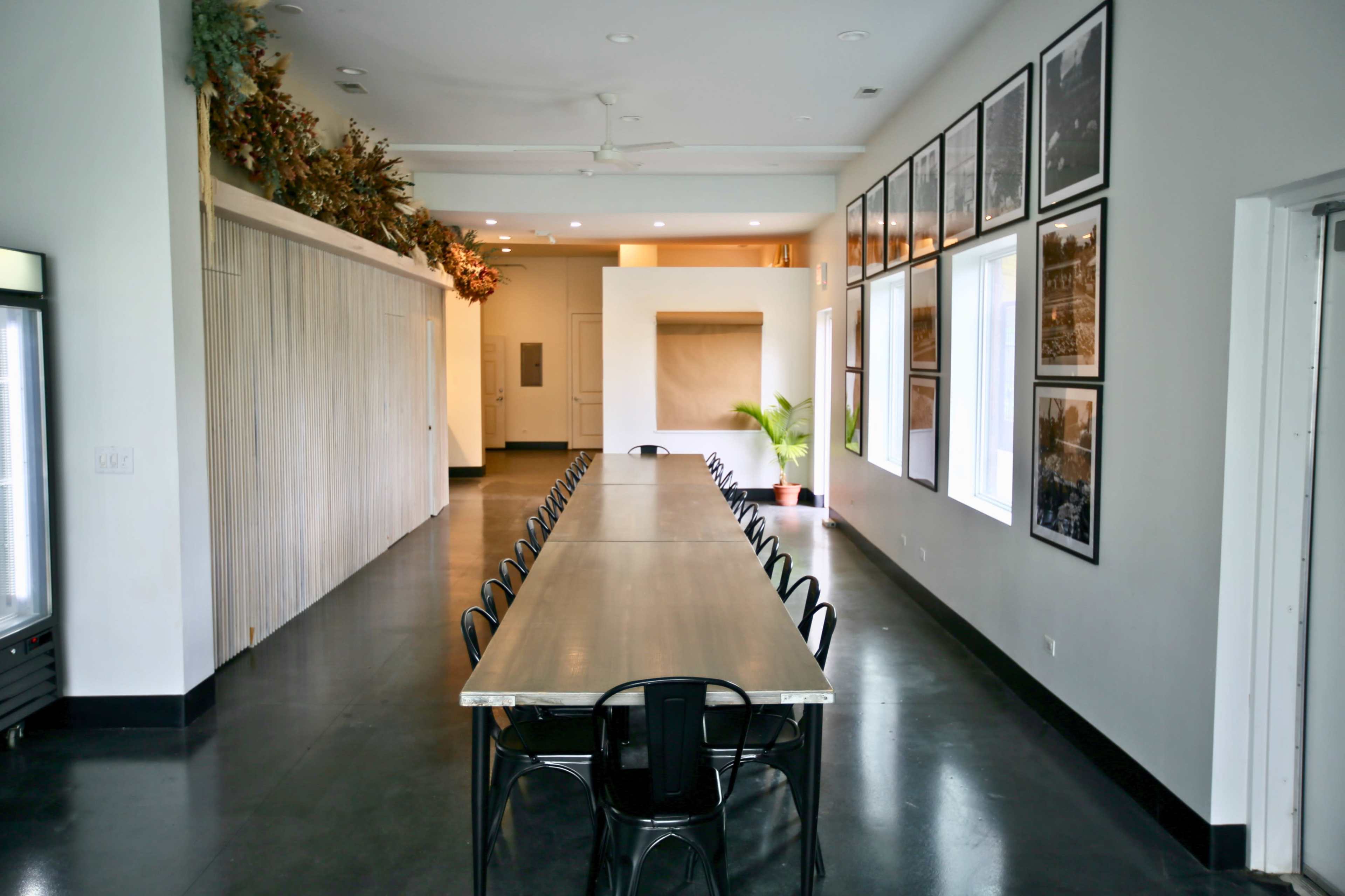 A long, rectangular dining table is placed in a spacious room with large windows and black flooring, surrounded by framed photographs on the walls.