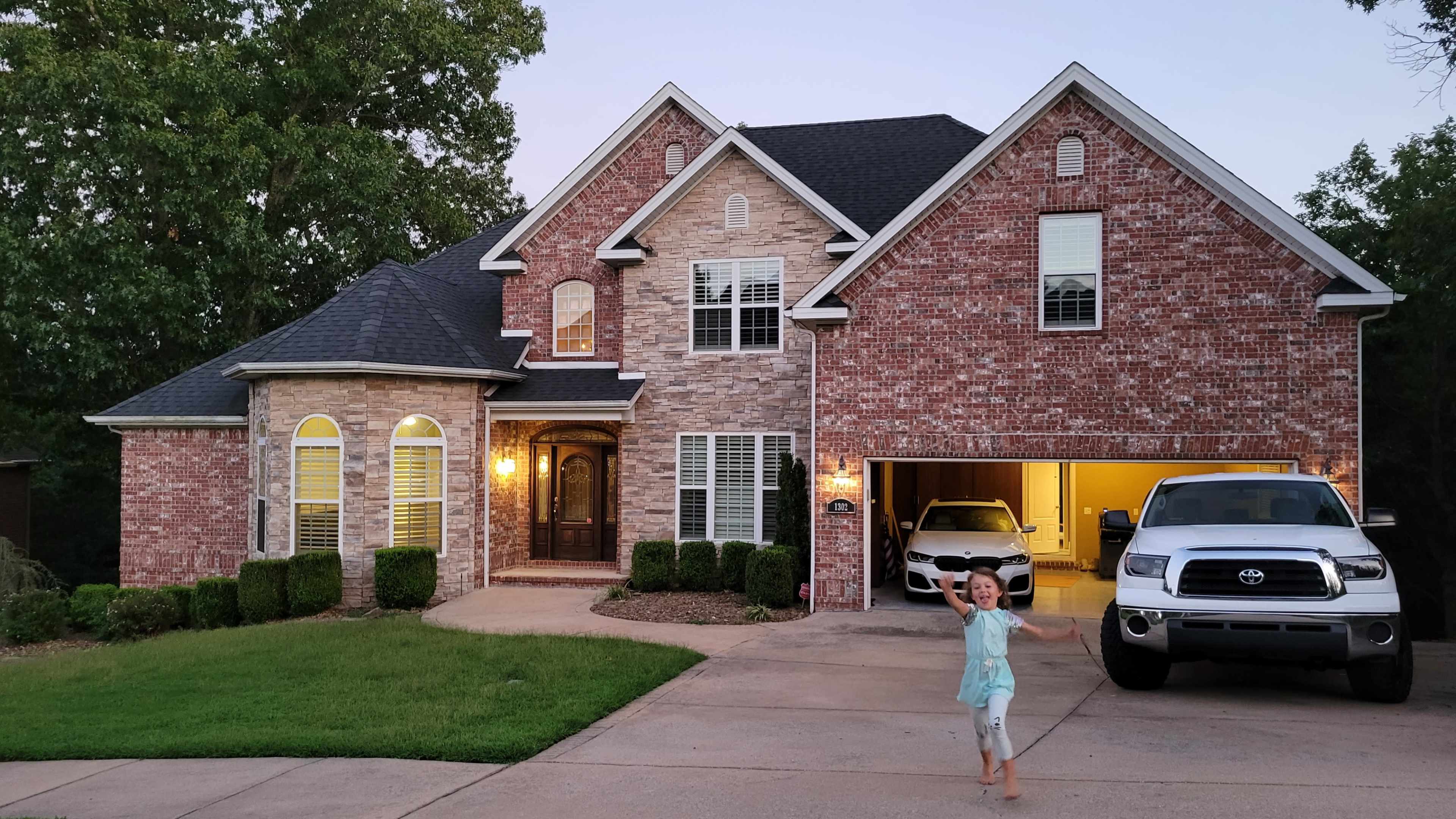 A child runs toward the camera in front of a large brick house with a neatly landscaped yard and a parked truck in the driveway.
