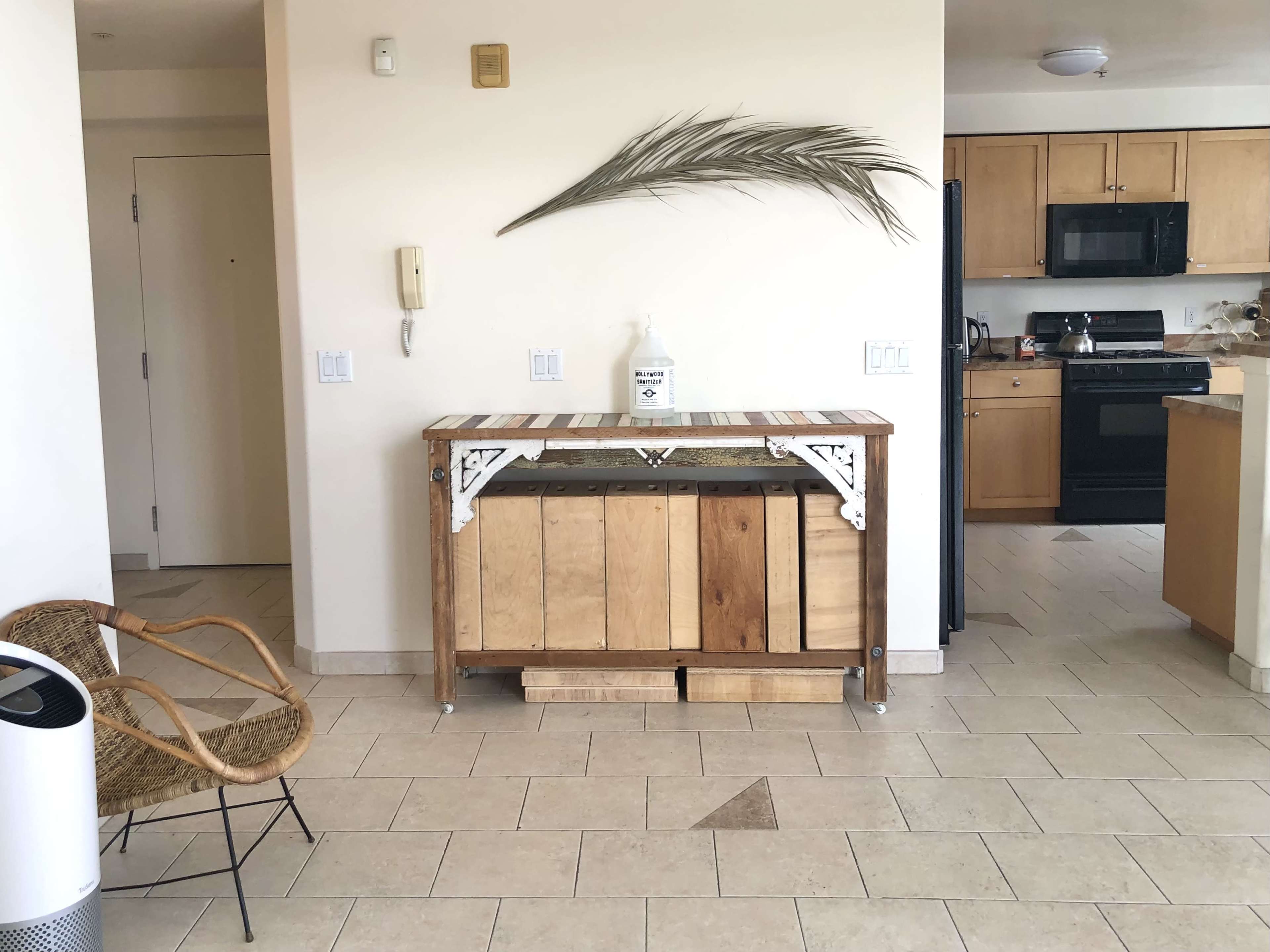 A wooden console table with a decorative palm leaf sits in an open space between a kitchen and a seating area.