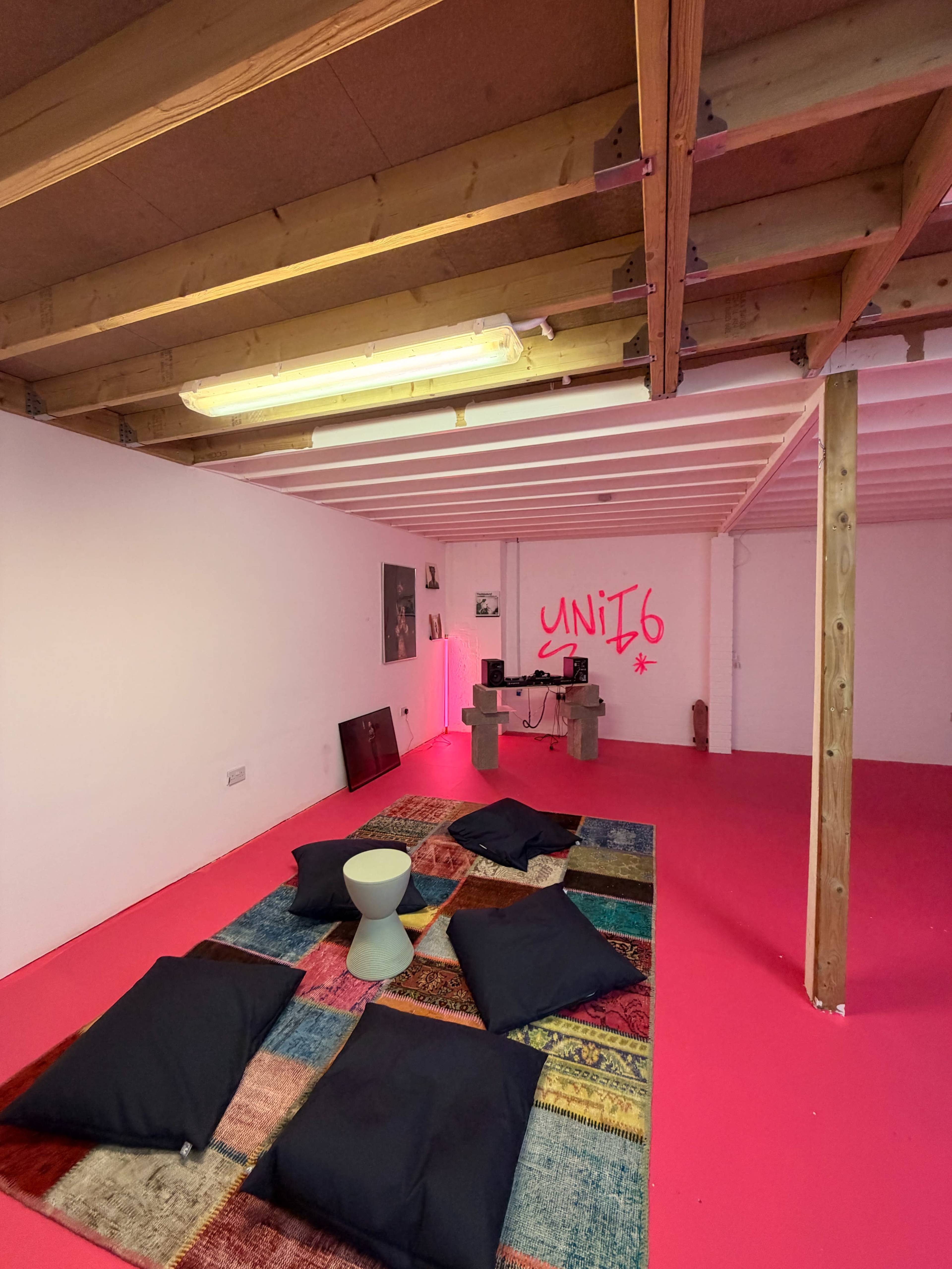The image depicts a brightly colored room with a pink floor, several large cushions arranged on a patterned rug, and a desk in the background with graffiti displaying "UNIT 6" on the wall.