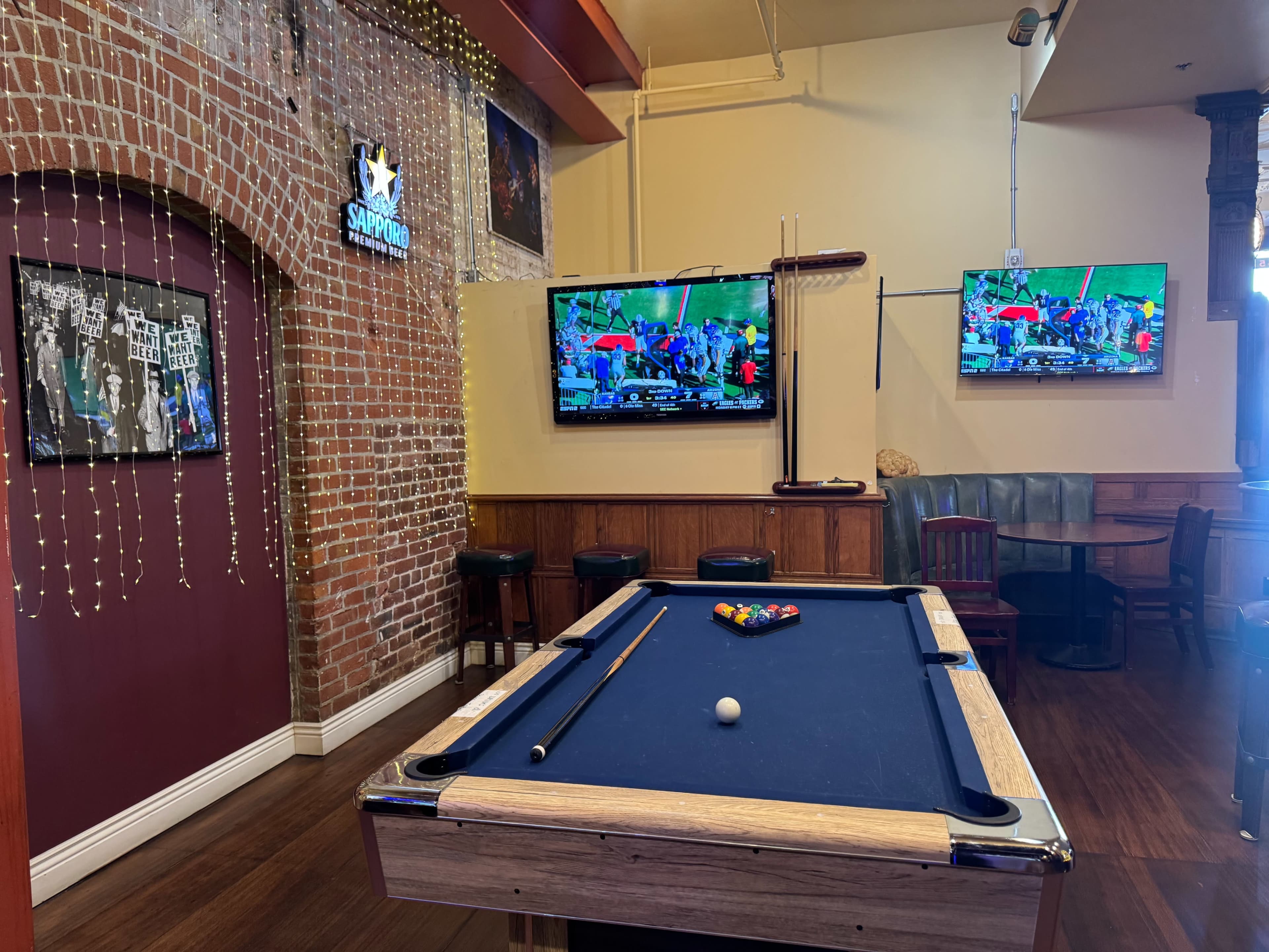The image shows a pool table in a recreational room with two televisions mounted on the wall, surrounded by brick and wooden finishes.