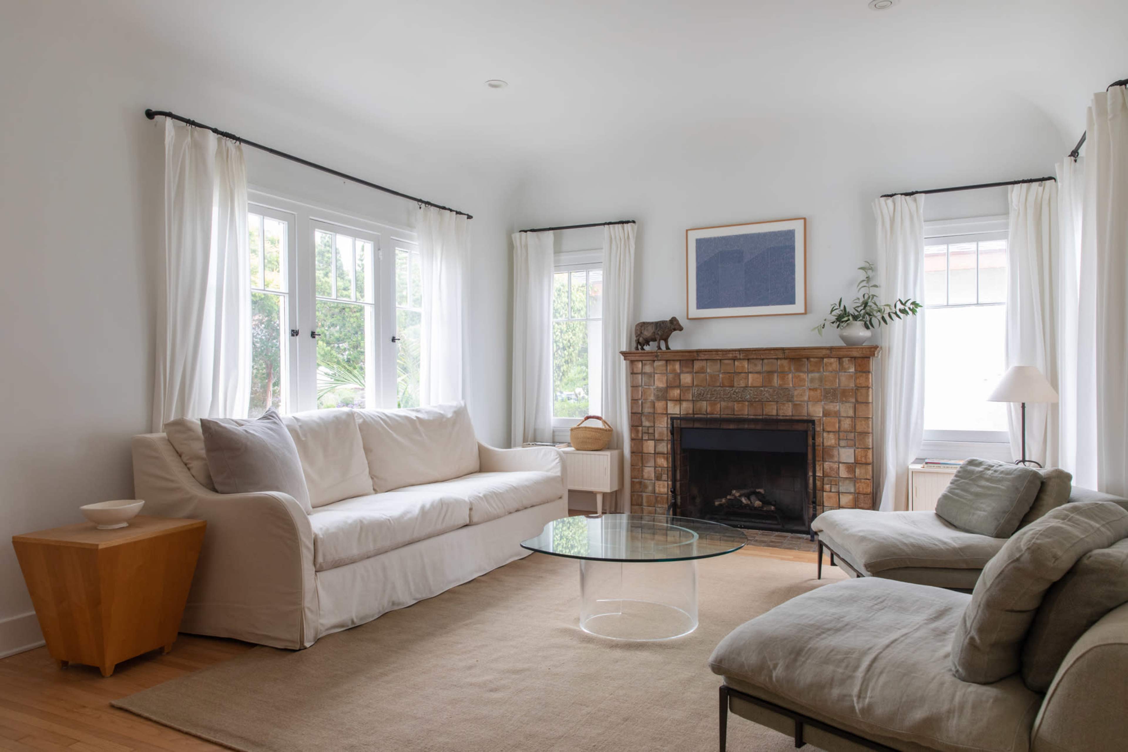 The living room features a white sofa, two light-colored armchairs, a glass coffee table, and a brick fireplace, all illuminated by natural light through large windows.