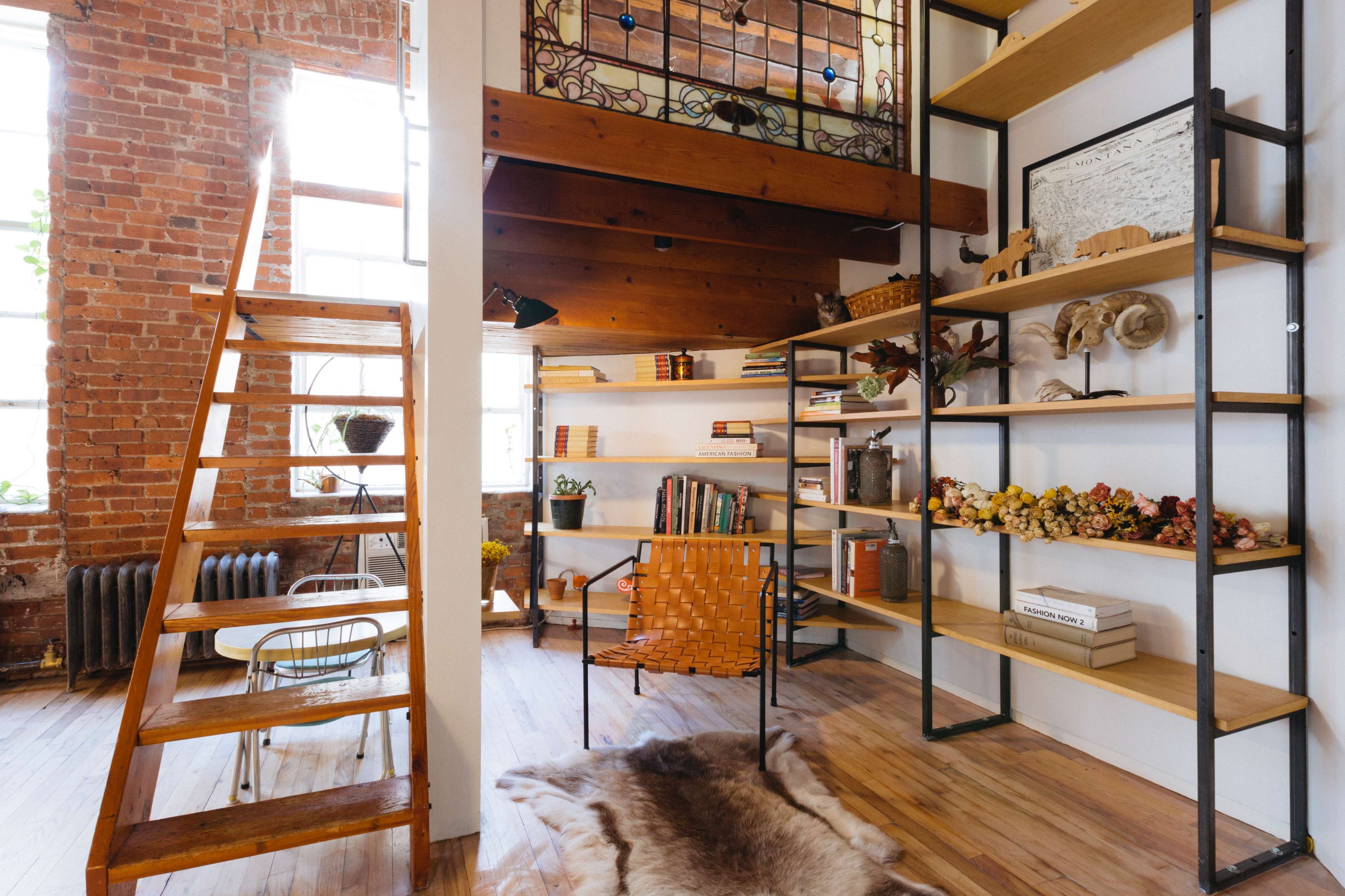 The image shows a cozy interior with wooden stairs leading to an upper level, a desk and chair arrangement beside open shelves filled with books and decorative items, and large windows allowing natural light to enter the space.