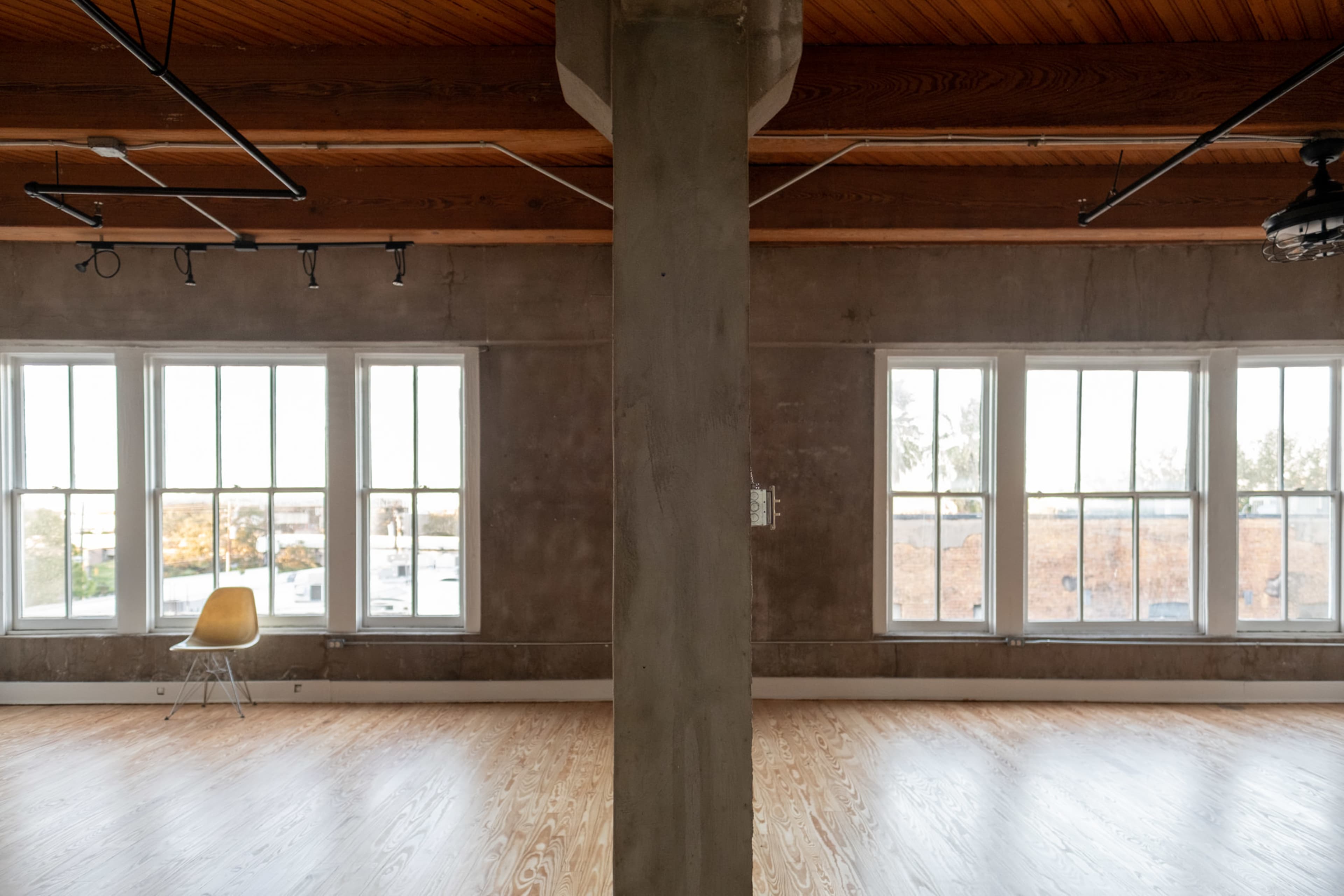 An empty interior space with a column in the center and large windows on either side, featuring a single chair positioned against one wall.