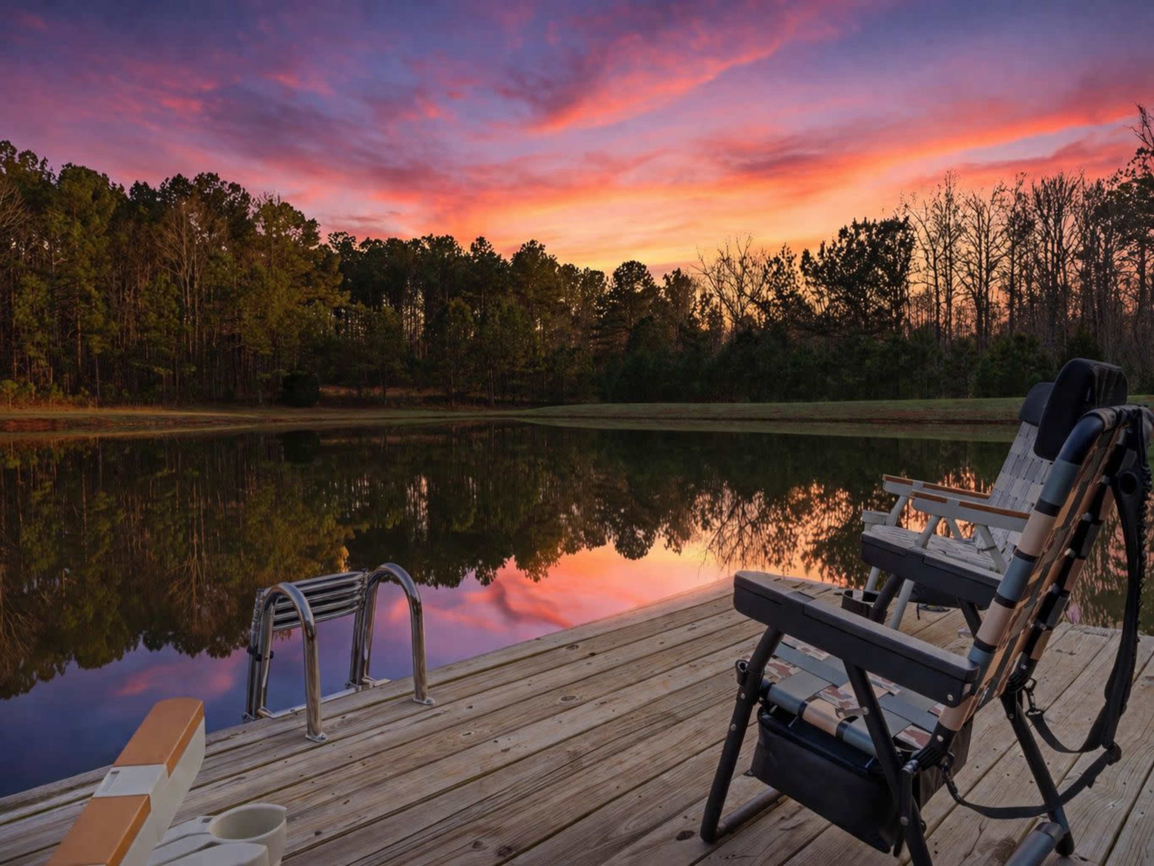 A wooden dock extends over a calm body of water, with two chairs positioned beside a small set of steps leading down, and the sky displaying vibrant colors at sunset.
