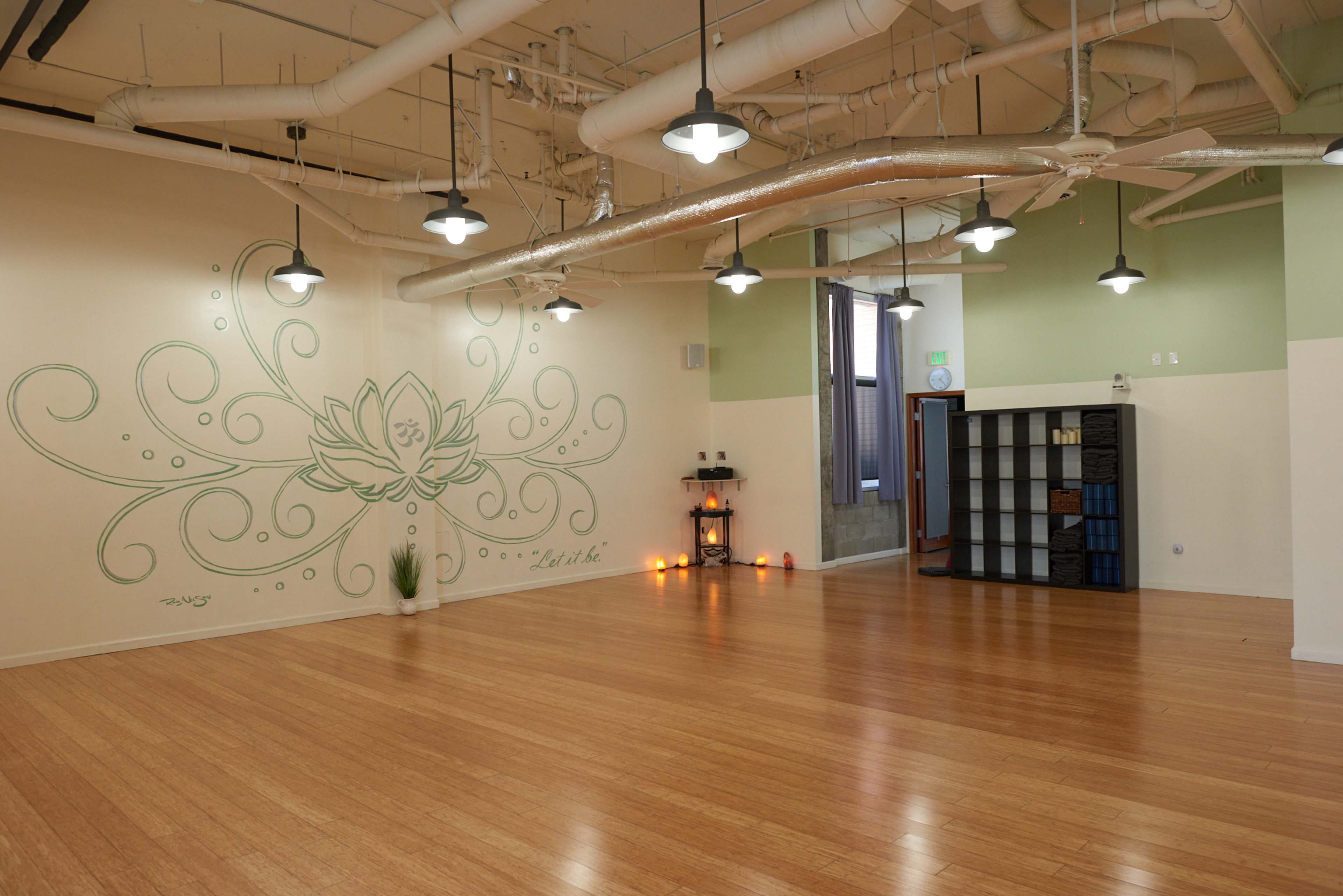 A spacious yoga studio with bamboo flooring, a green wall mural of a lotus flower, and various decorative elements.