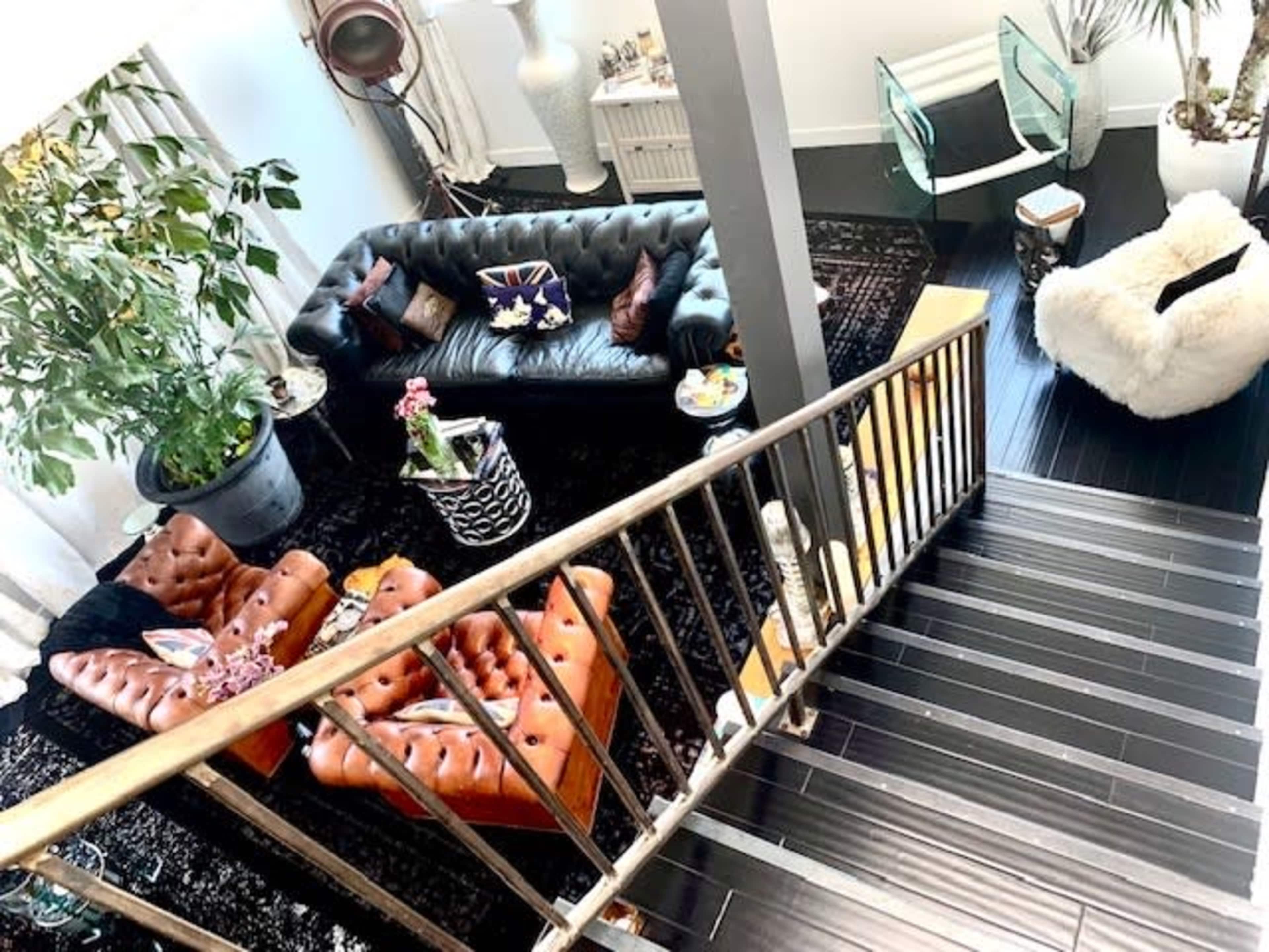 A stylish living room with a black leather sofa, brown tufted armchairs, a large plant, and a staircase leading to an upper level.