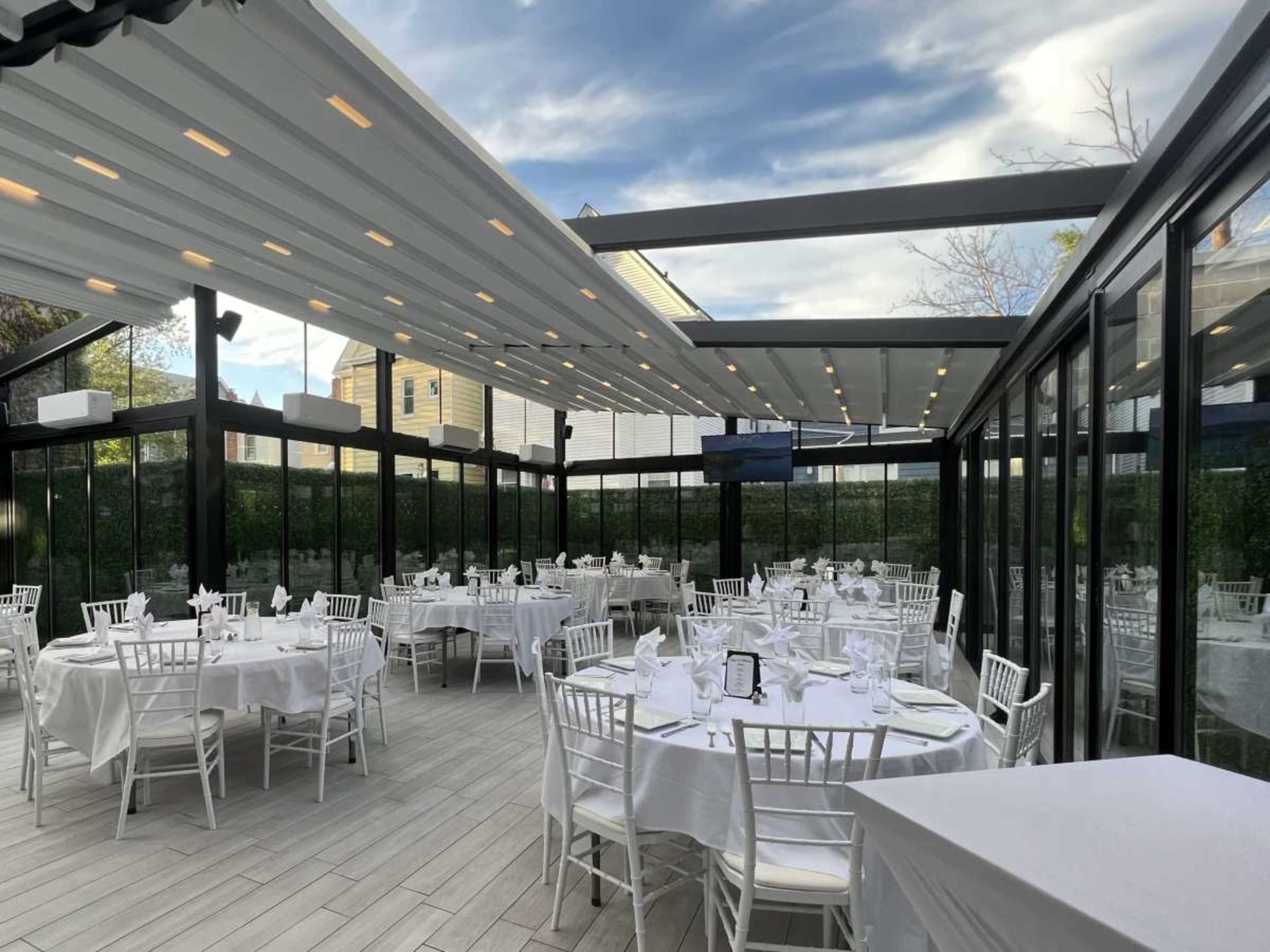 The image shows an elegant dining area with white tables set for a formal event, enclosed by large glass panels.