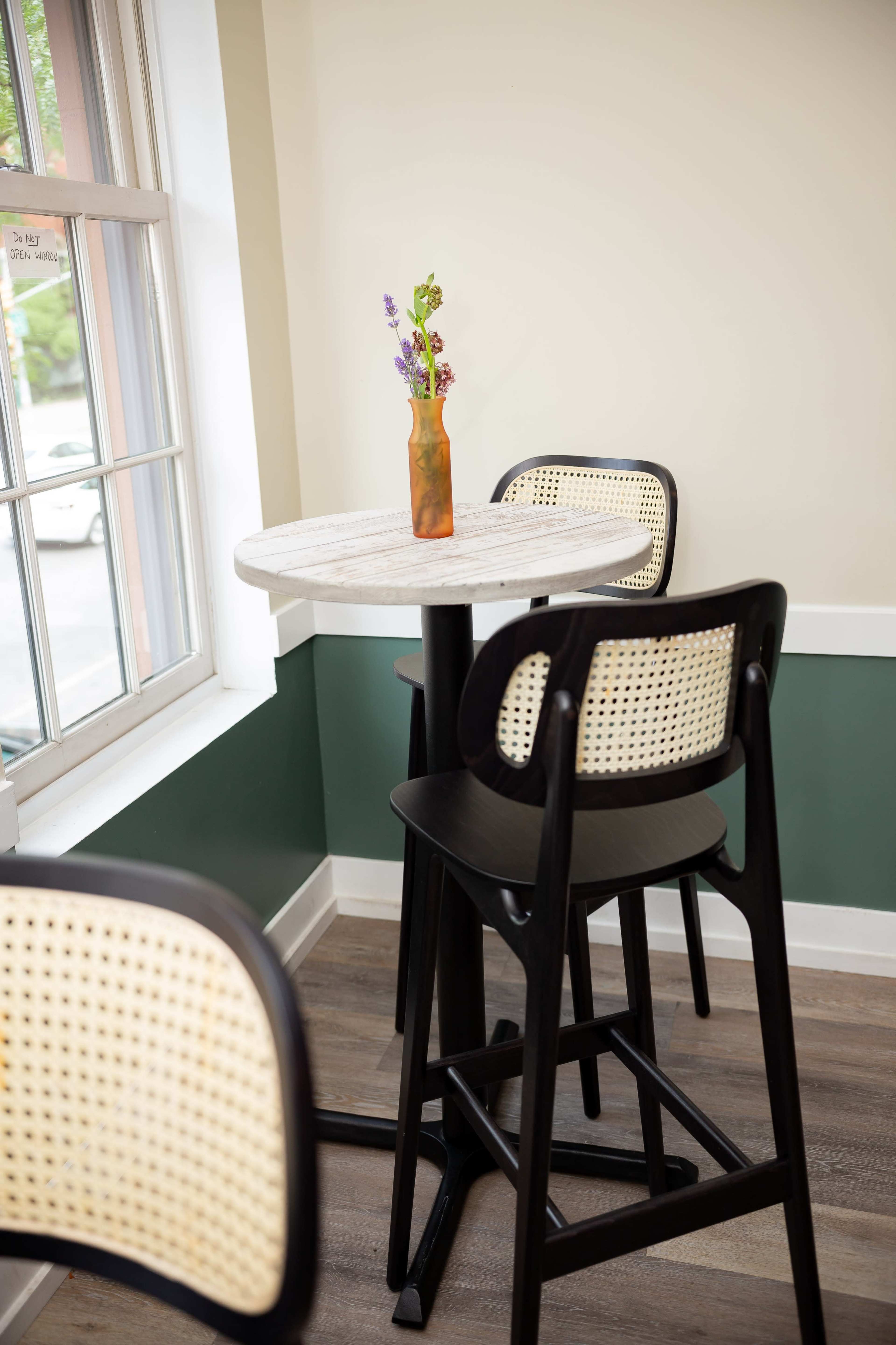 A small round table with a decorative vase containing flowers is positioned between two high-backed chairs near a window.