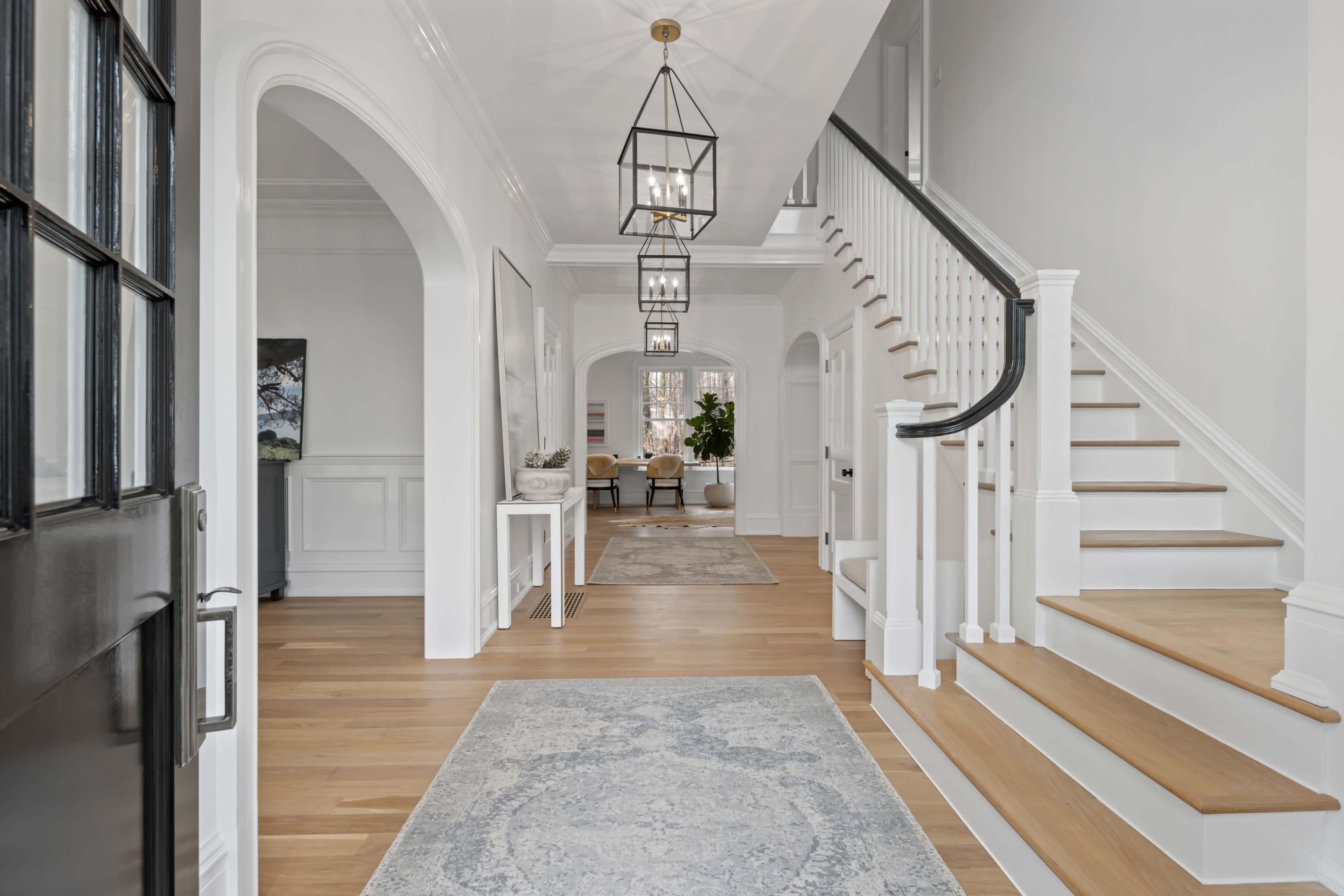 A bright foyer features a staircase, modern light fixtures, and a table beside an archway leading to a sitting area.